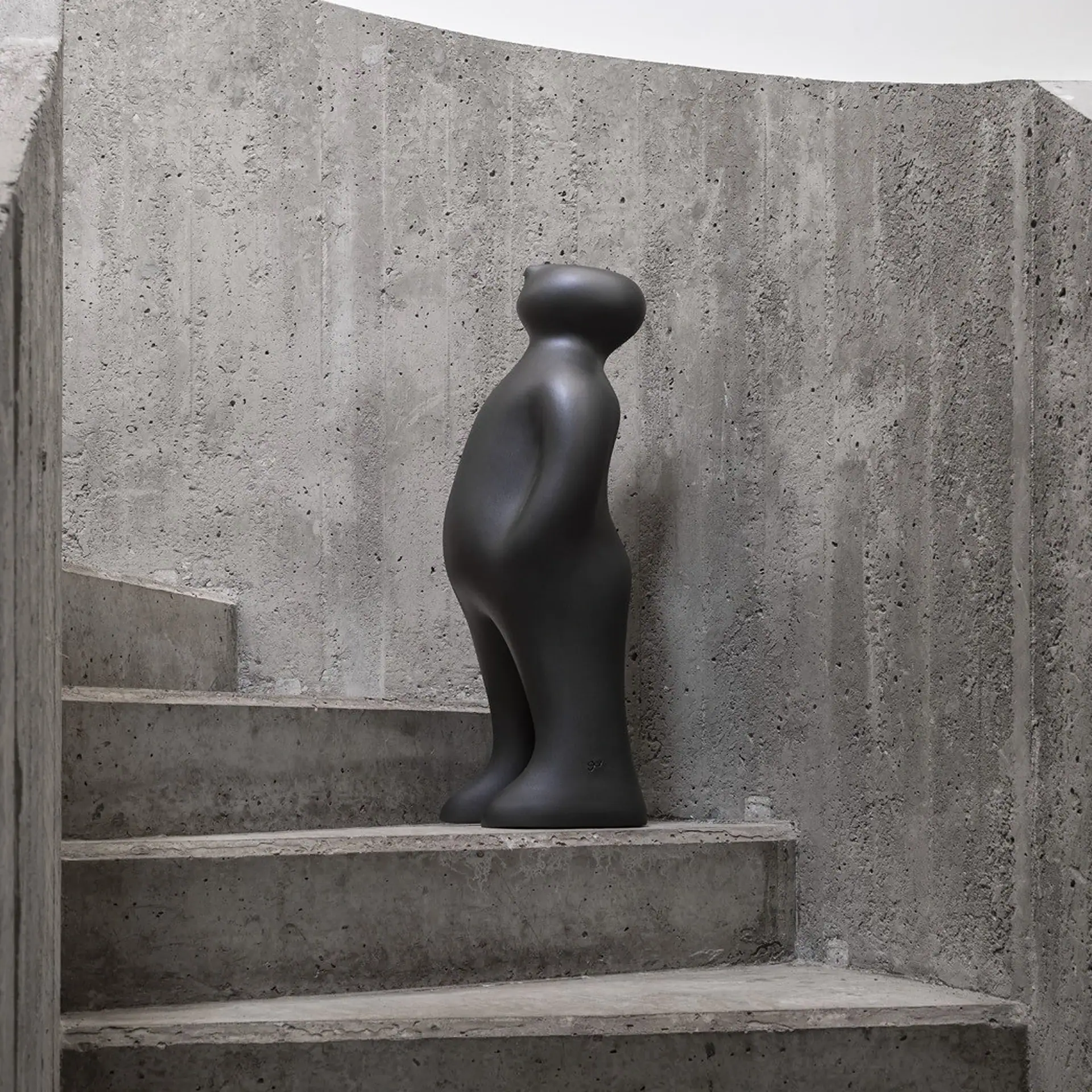 A dark grey, abstract, human-like sculpture with a smooth, matte finish stands on a rough, curved concrete staircase.