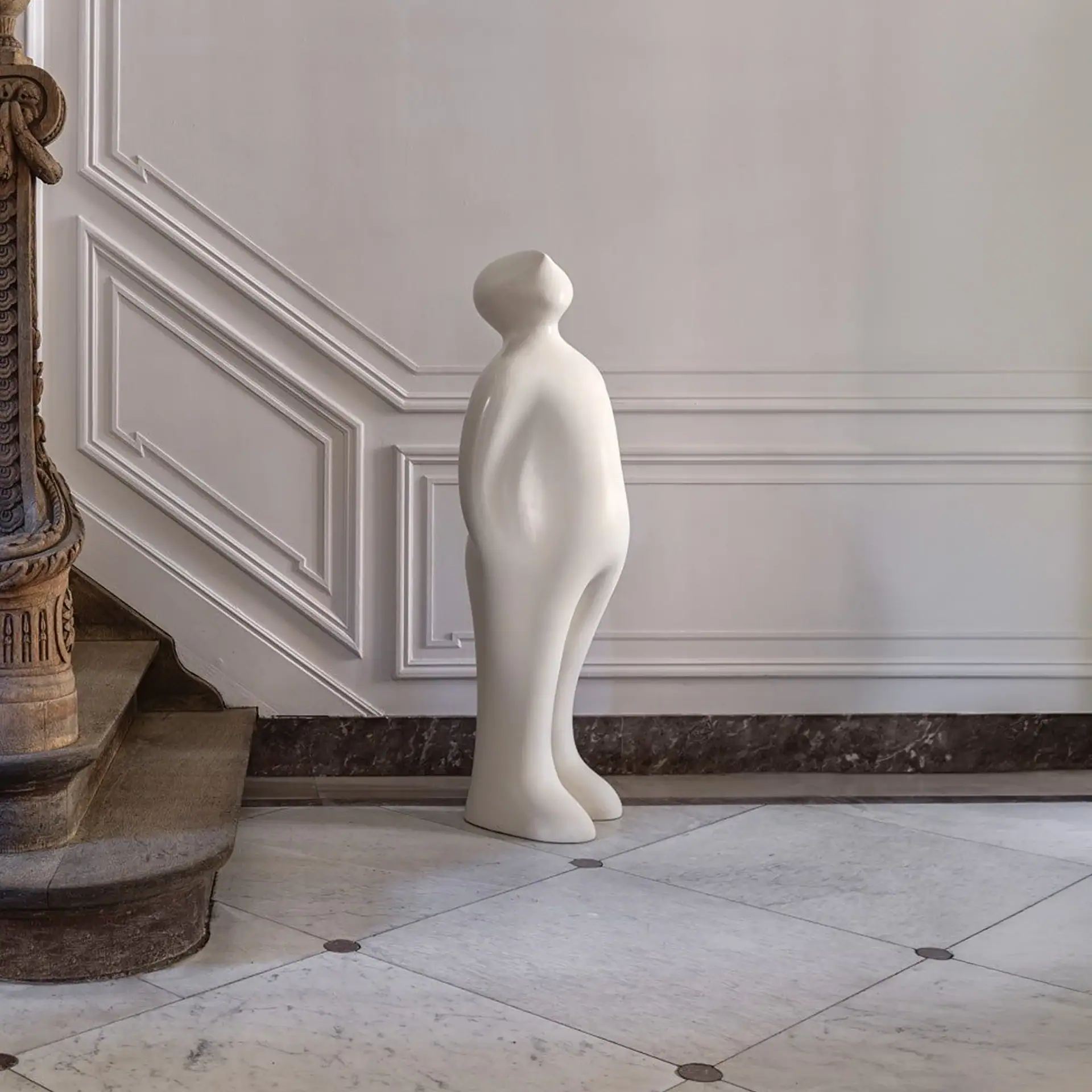 A smooth, white abstract human sculpture stands on a marble floor in front of a paneled wall and an ornate wooden staircase.