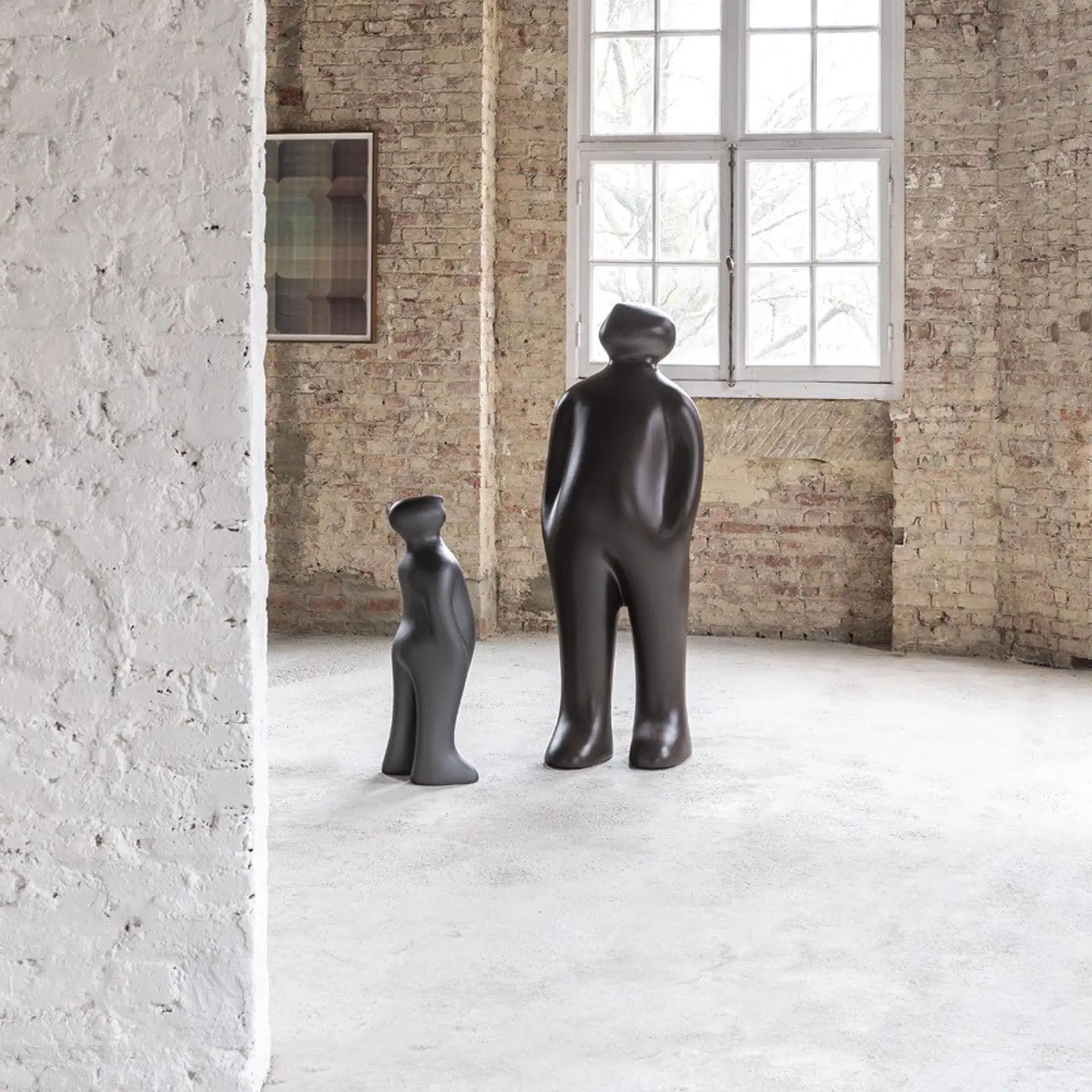 Two abstract, human-like sculptures, one large dark brown and one smaller gray, stand in an industrial room with brick walls and a large window.