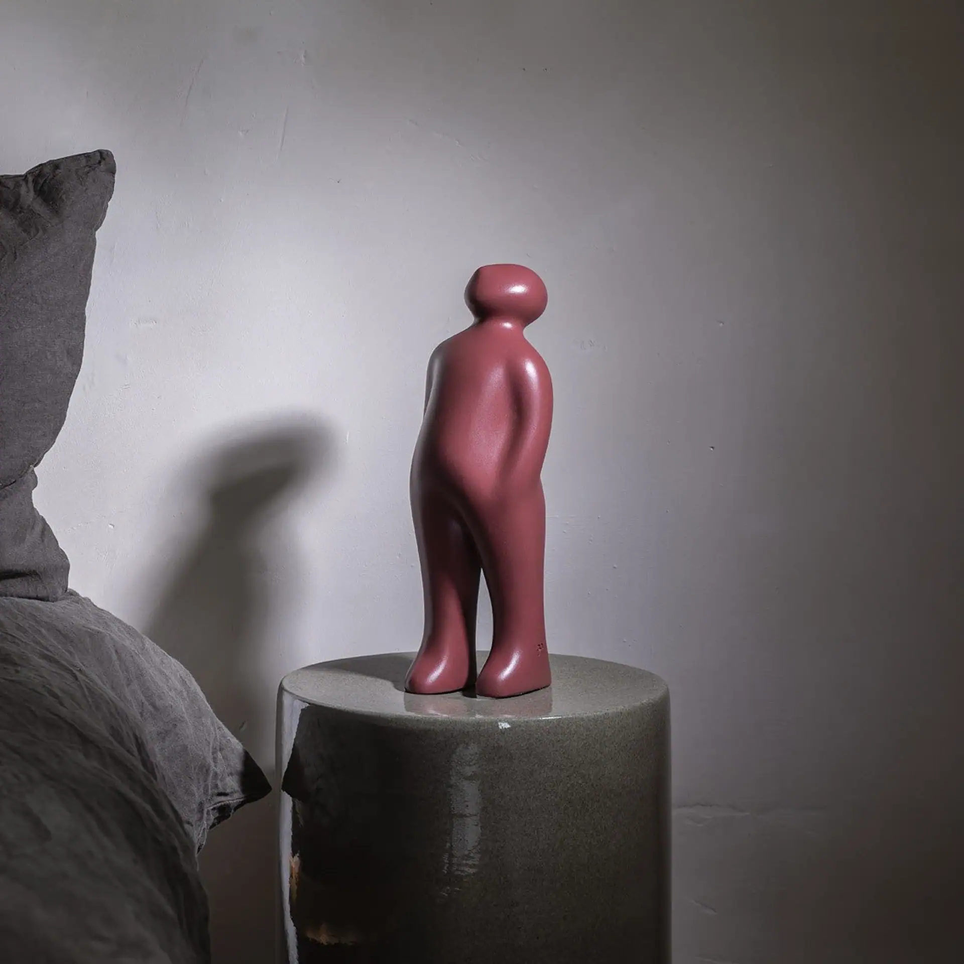 A reddish-pink abstract human-like figurine on a dark gray cylindrical pedestal, casting a long shadow on a light wall next to dark gray fabric.