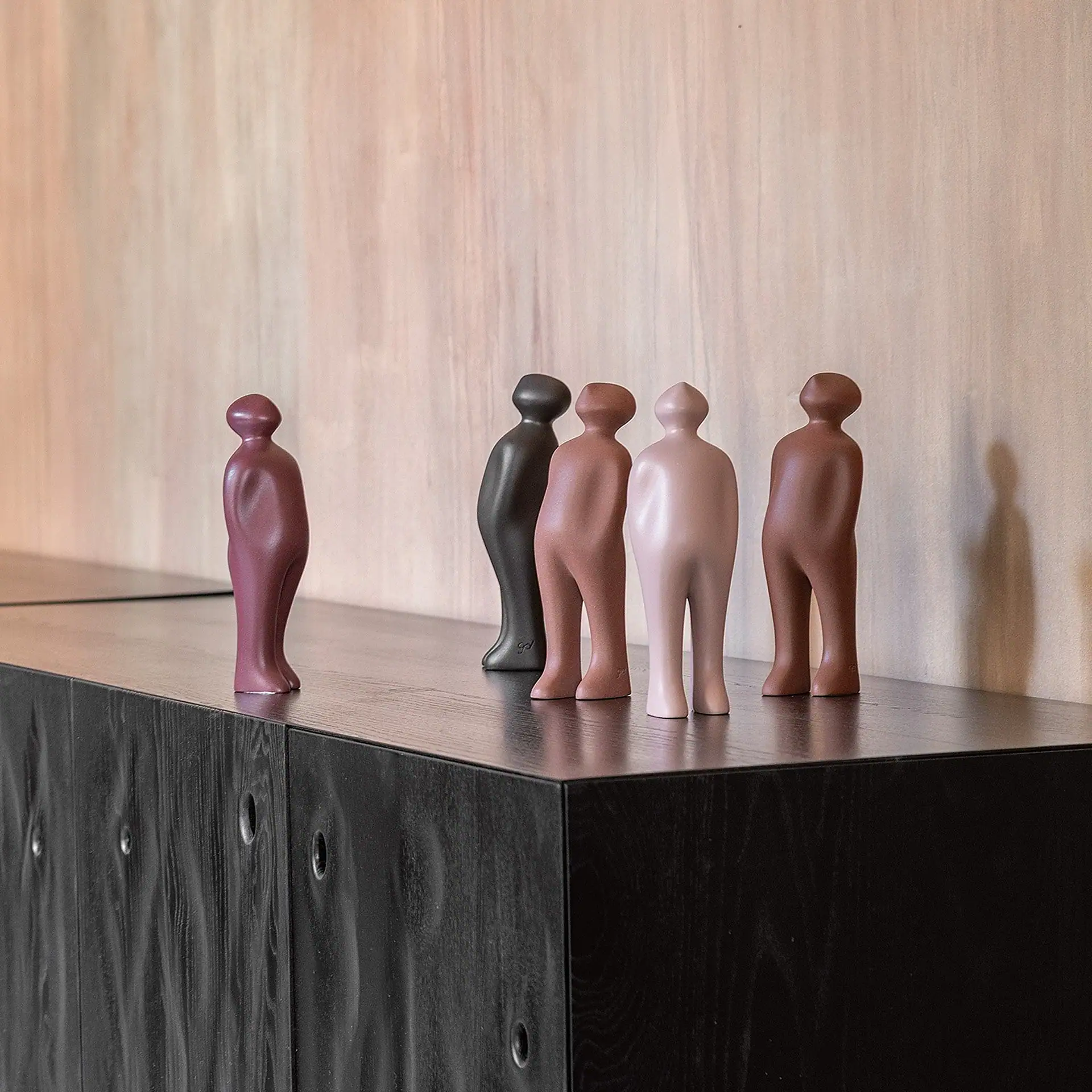 Five abstract human-shaped figurines in maroon, dark gray, reddish-brown, light pink, and medium brown colors stand on a dark wooden cabinet against a light textured wall.