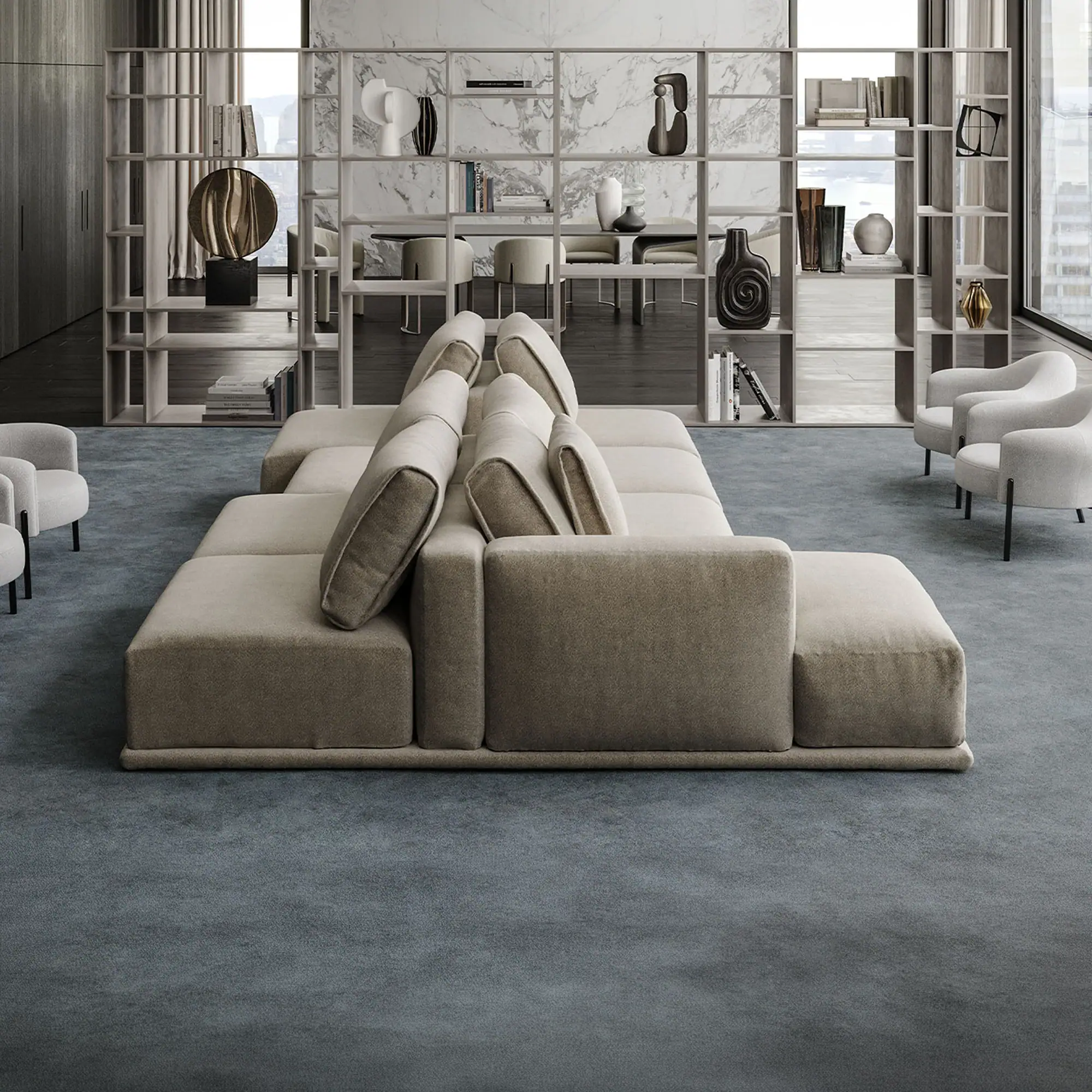 A large, light beige modular sofa on a blue-grey carpet in a modern living space with an open shelving unit and white armchairs.