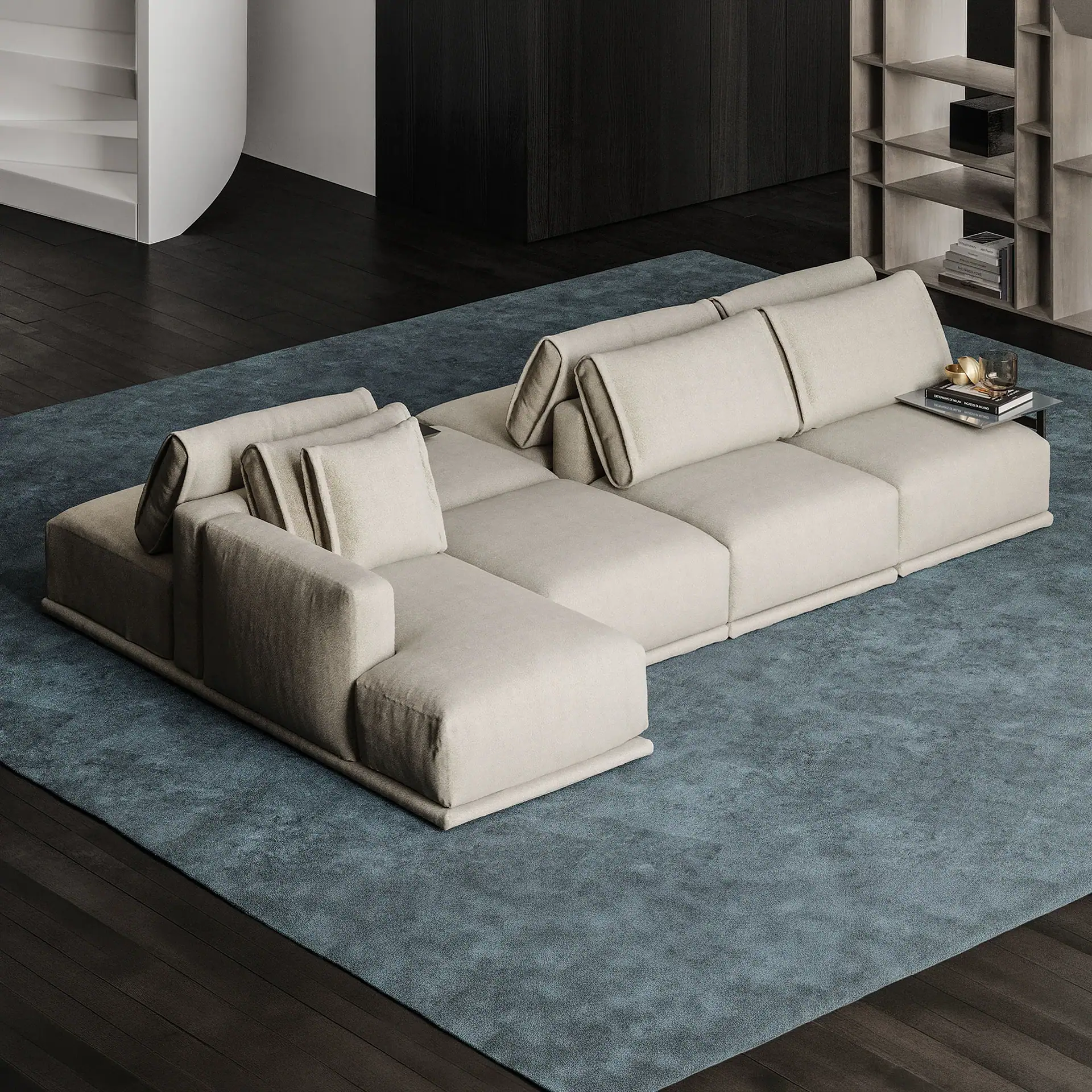 A modern L-shaped light beige sectional sofa on a blue rug, with dark wooden floors, a white curved staircase, and wooden shelving in the background.