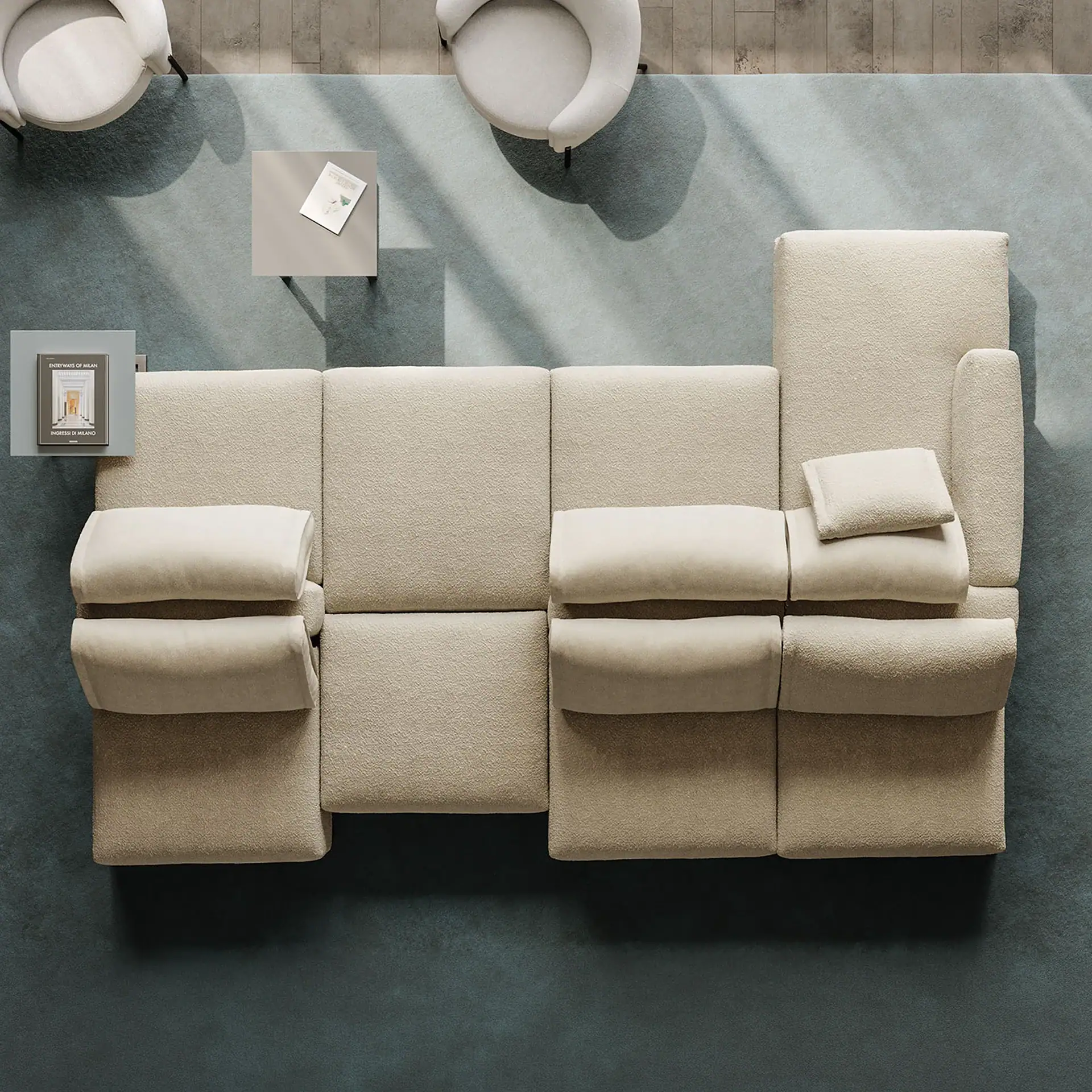 Overhead view of a modern living room with a light beige modular sofa, two armchairs, and two side tables on a blue-grey rug over a wooden floor. Natural light casts shadows.