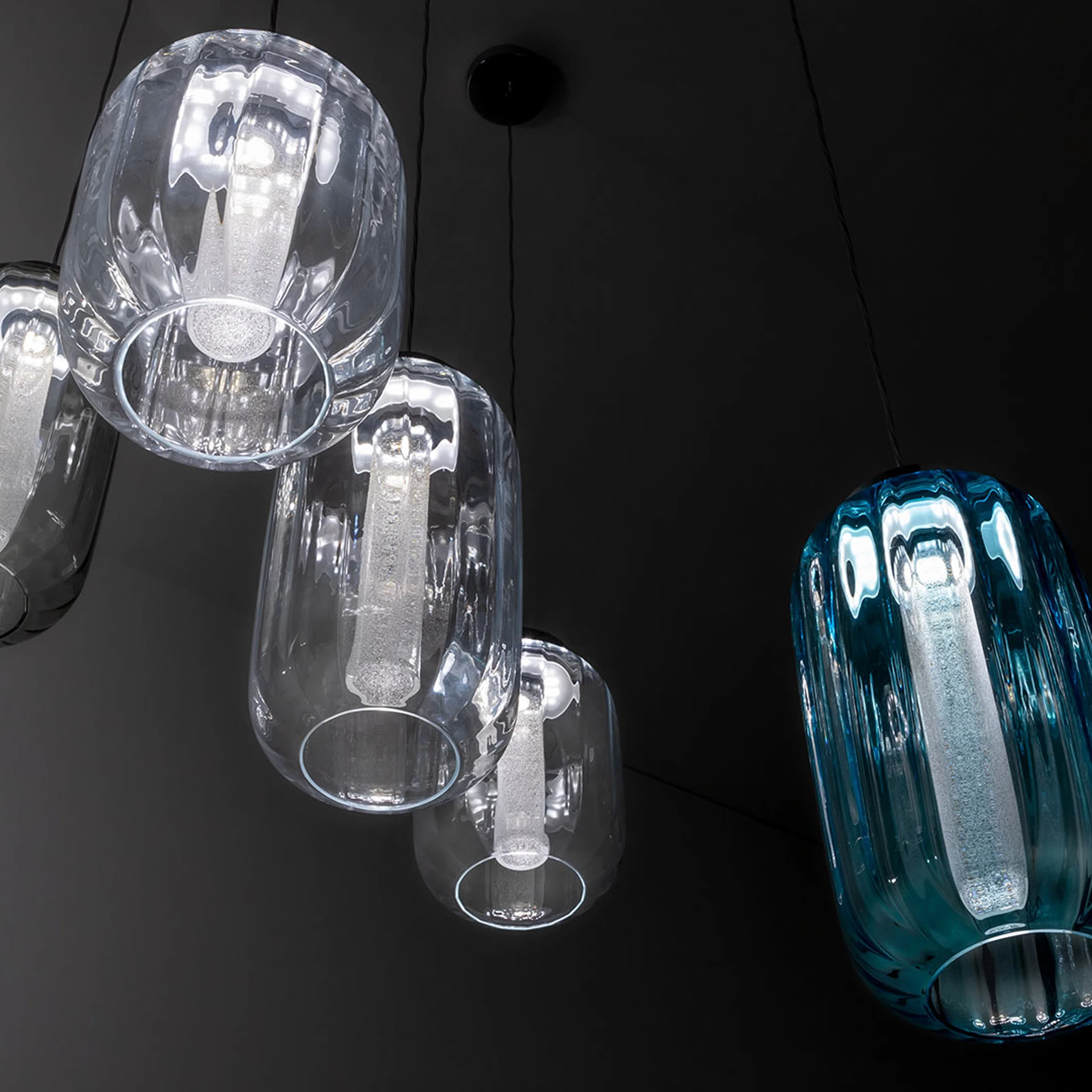 Five pendant lights hanging from a dark ceiling, four clear glass and one blue glass, all with ribbed shades and illuminated cylindrical bulbs.
