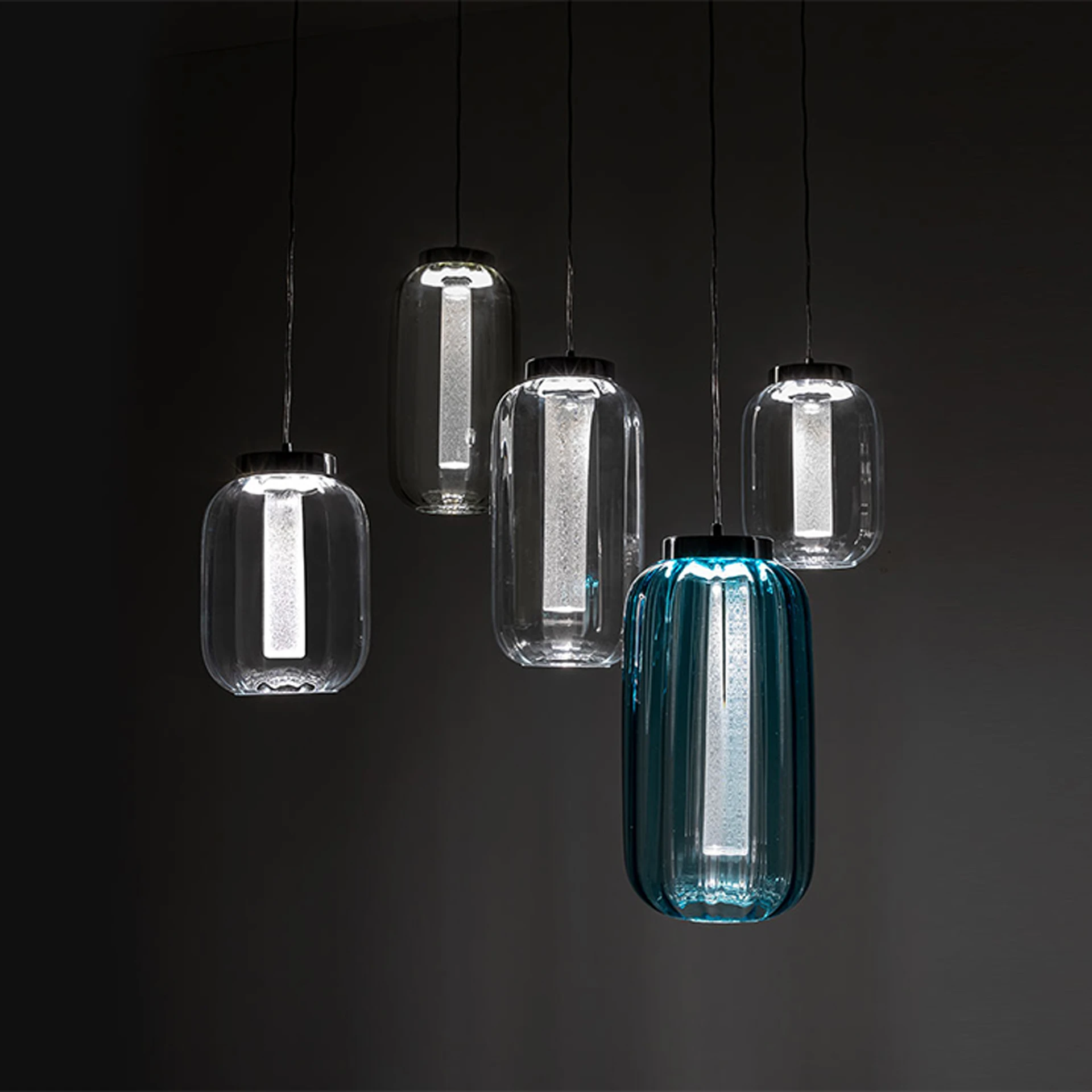 Five modern pendant lights hanging against a dark background. Four are clear glass, one is teal-blue ribbed glass, all with internal white light sources.