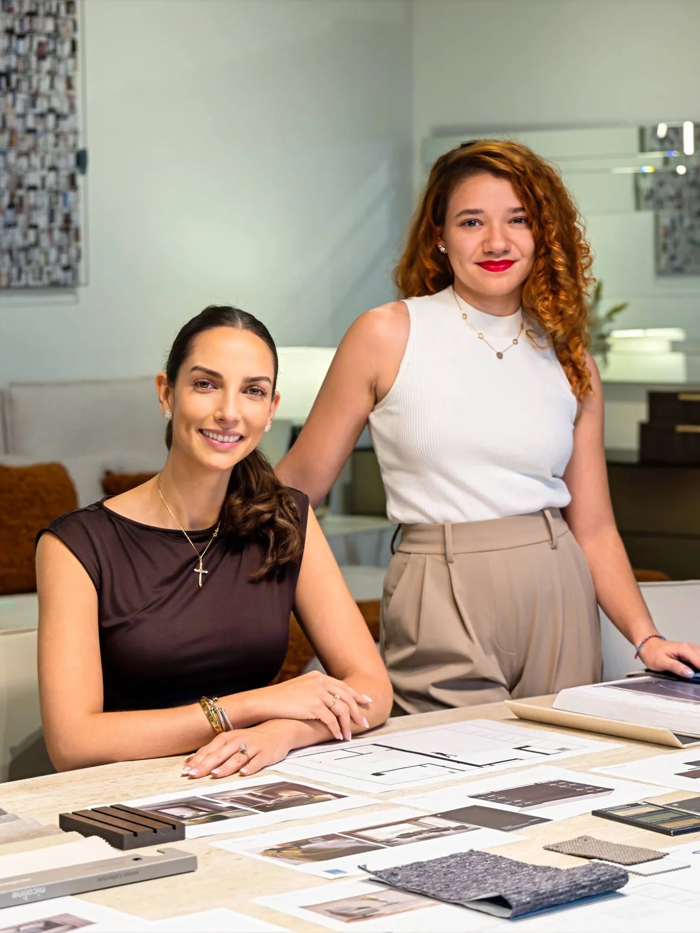 Two smiling women in a design studio, one seated at a table with design materials, the other standing behind her.
