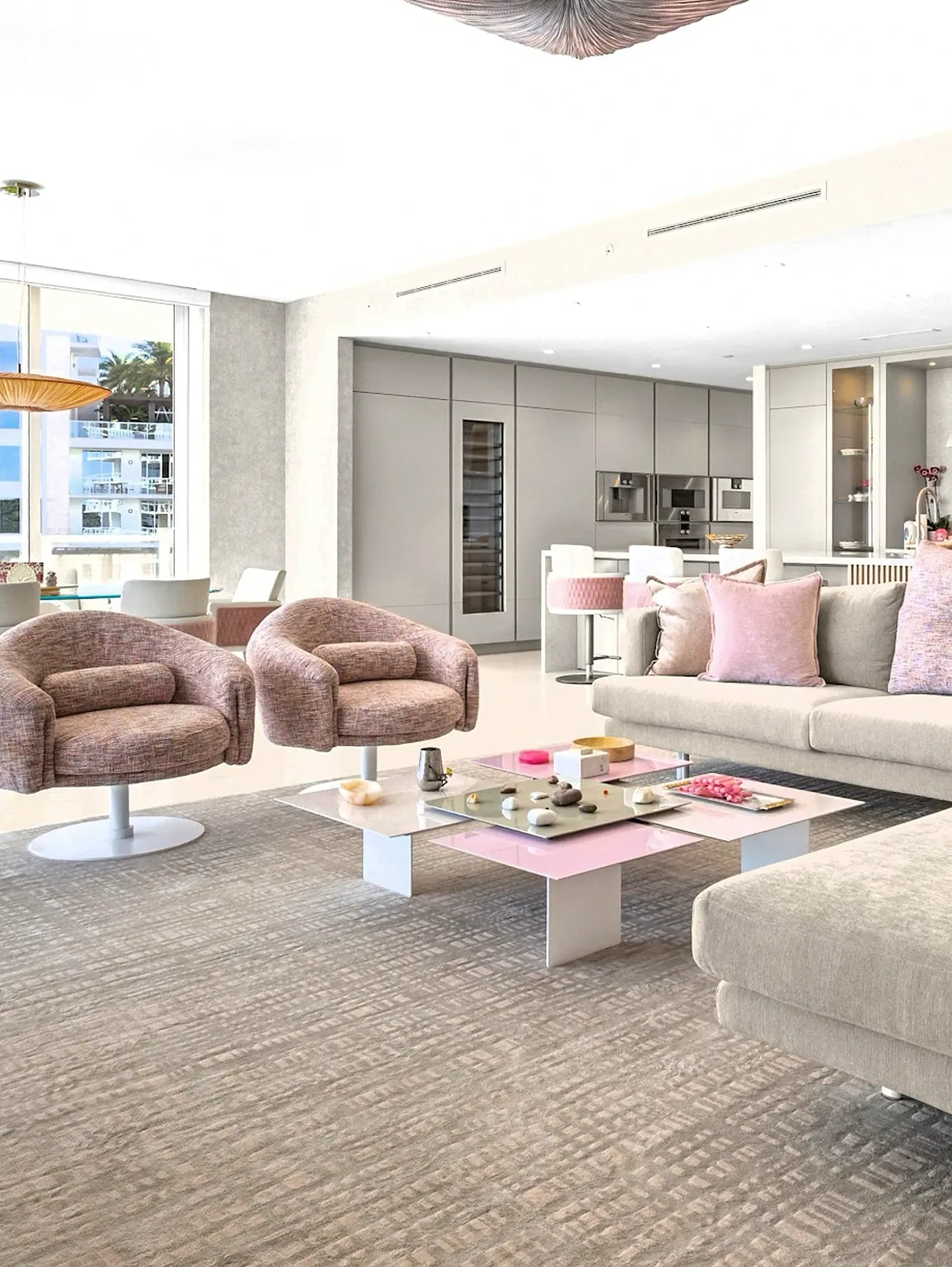 A modern open-concept living room and kitchen with pink and neutral decor, featuring a sofa, armchairs, coffee table, and a view of a city through a large window.
