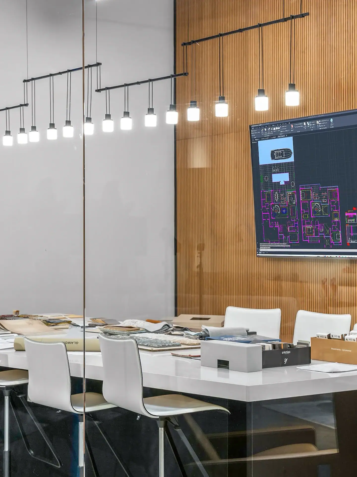 Interior of a modern design studio with a white table, chairs, design material samples, and a large monitor displaying architectural CAD plans.