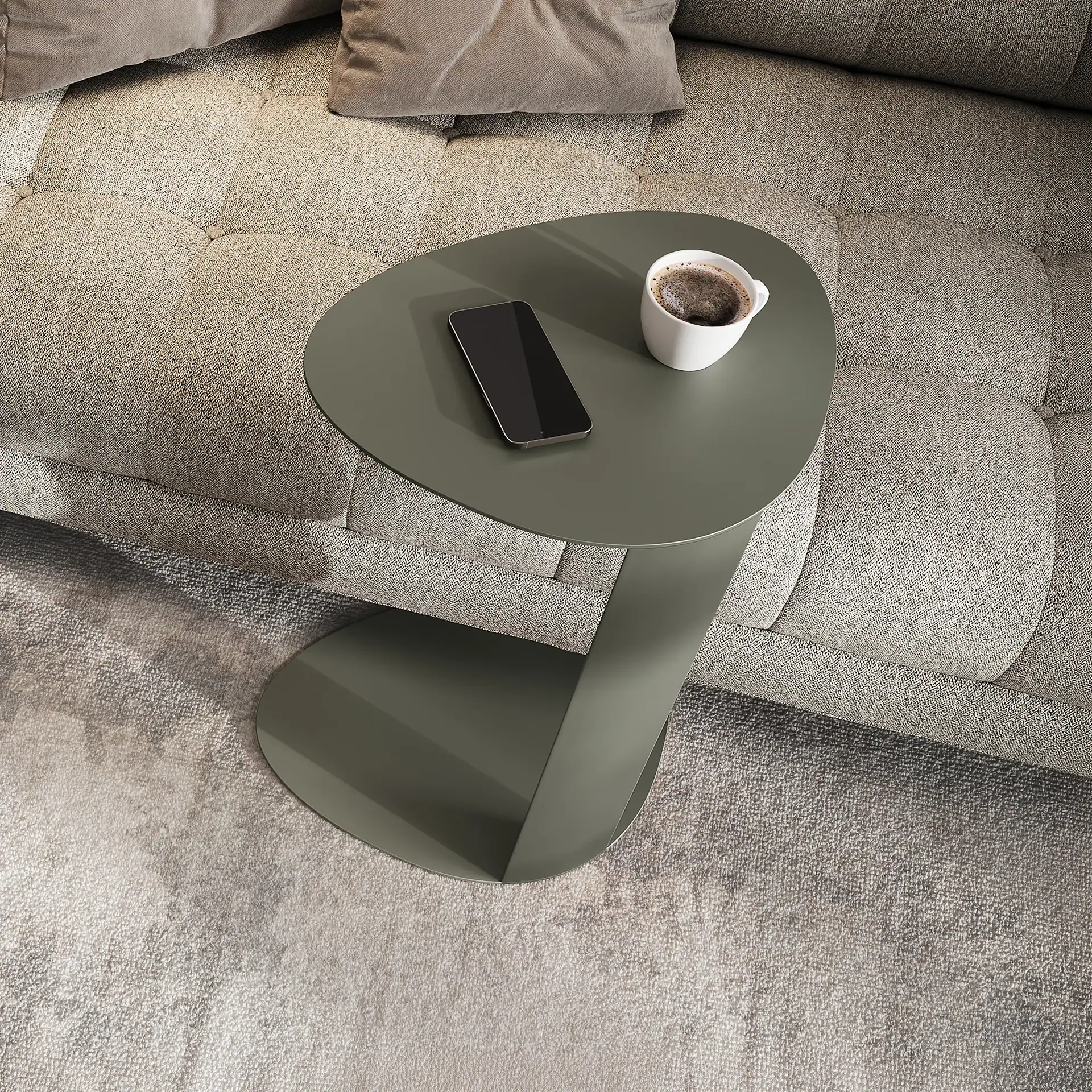 Overhead view of a dark green C-shaped side table with a white coffee mug and a black smartphone, next to a light grey tufted sofa on a textured rug.
