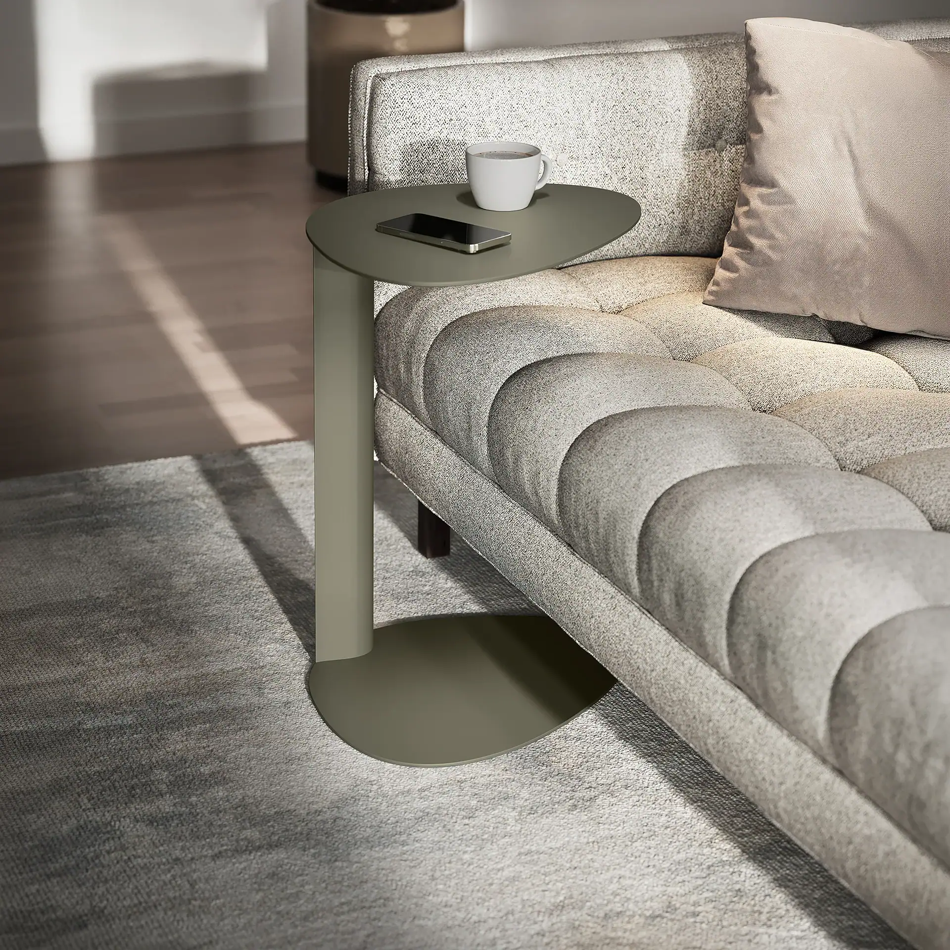 A sage green side table with a coffee mug and smartphone next to a light grey tufted sofa.