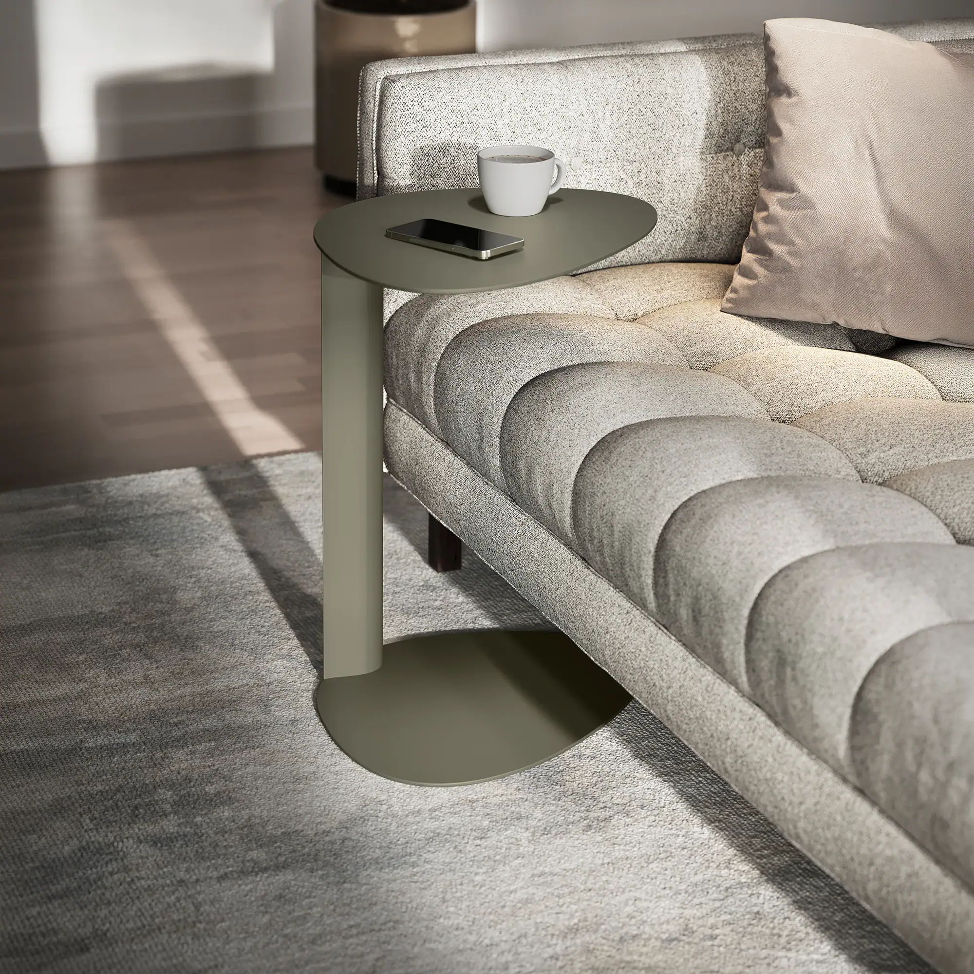 A sage green side table with a coffee mug and smartphone next to a light grey tufted sofa.