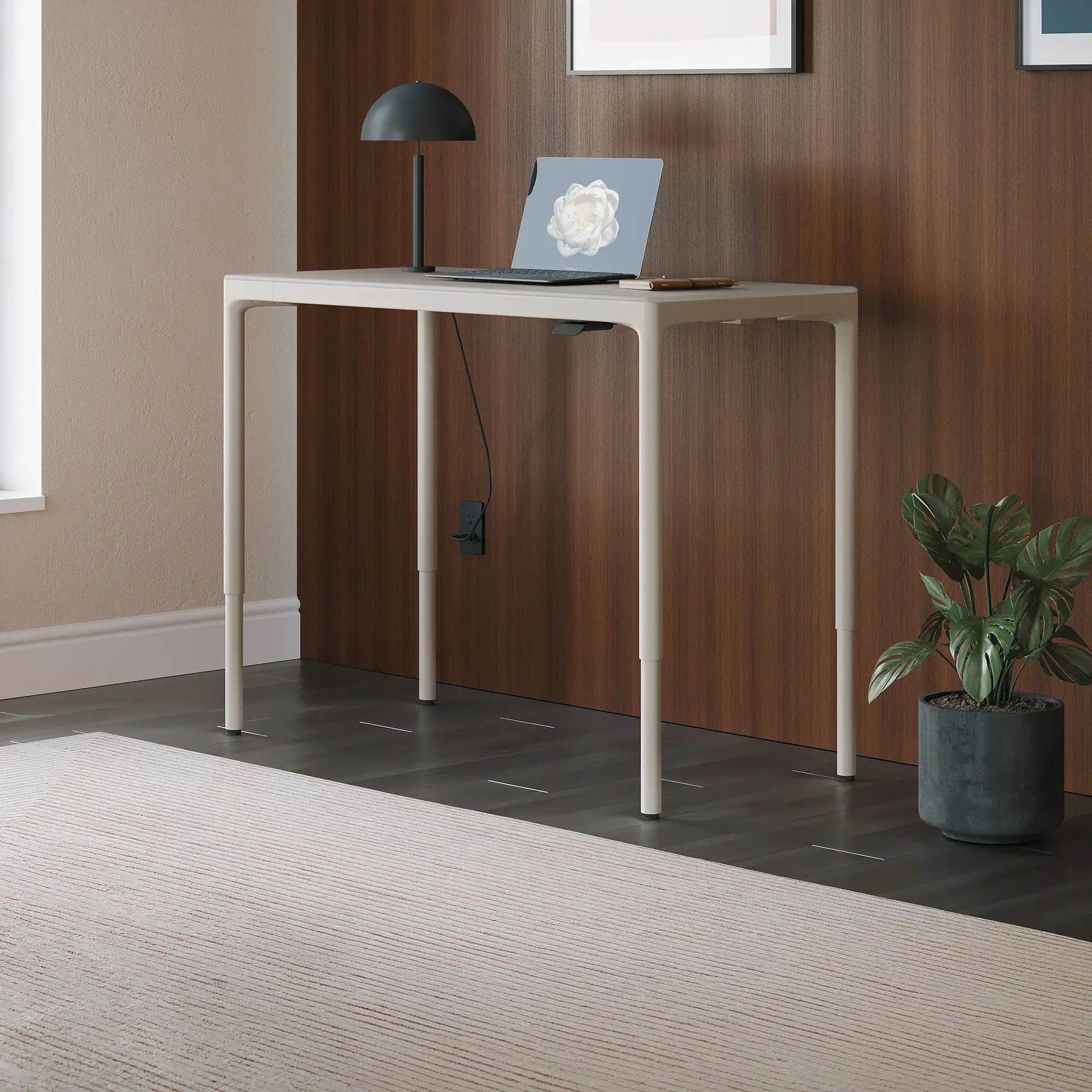 A light beige standing desk with a laptop and lamp, a Monstera plant, and a striped rug in a modern room with wood paneling and a beige wall.