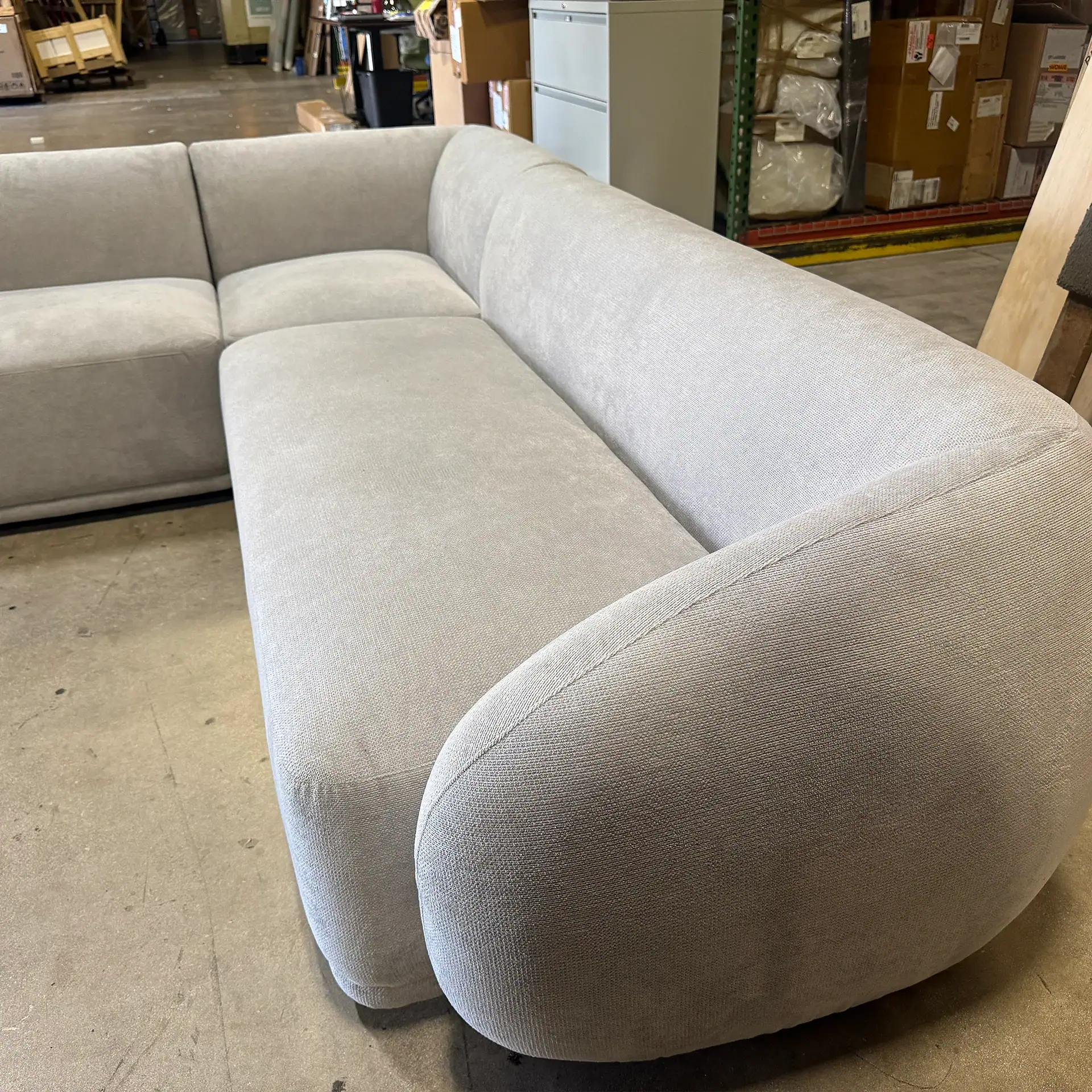 Modern light gray sectional sofa with rounded armrest in warehouse setting.