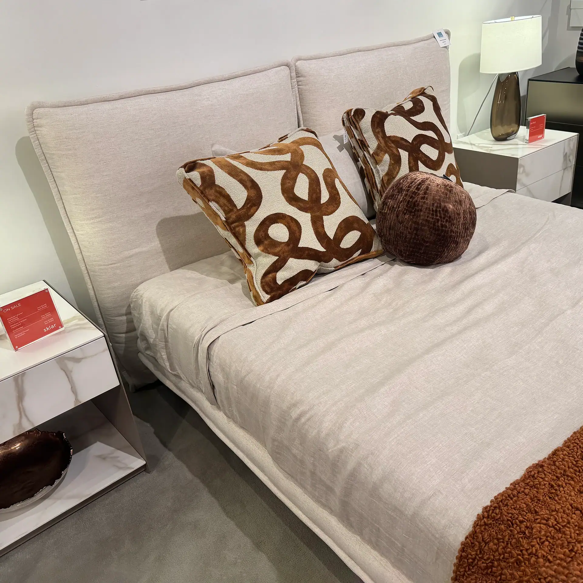 Queen bed with beige upholstery and decorative pillows in a modern bedroom setting.
