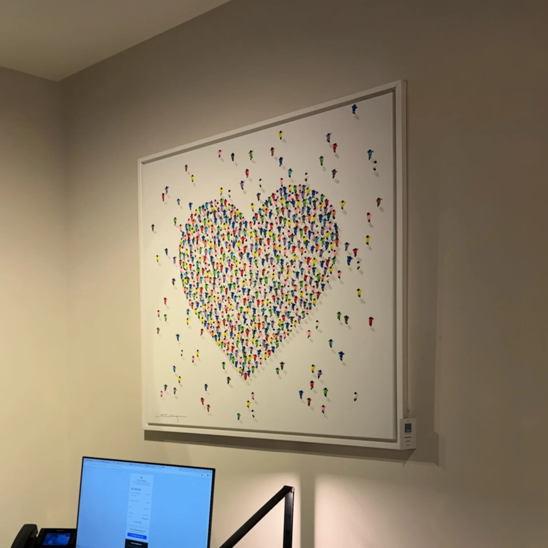 A large framed artwork on a light beige wall, depicting a heart shape made of many small, colorful human figures on a white background. A computer monitor and desk phone are visible below.