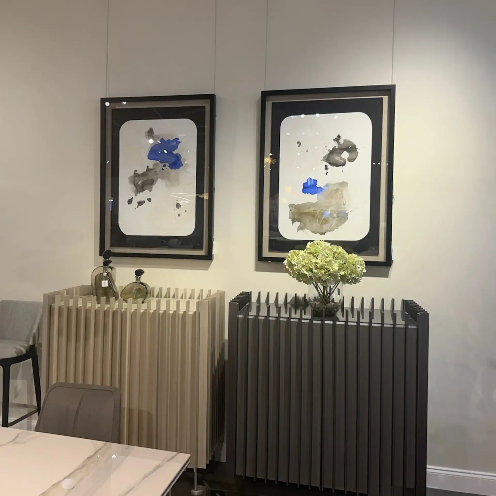 Two framed abstract paper paintings on a wall with decorative plants below.