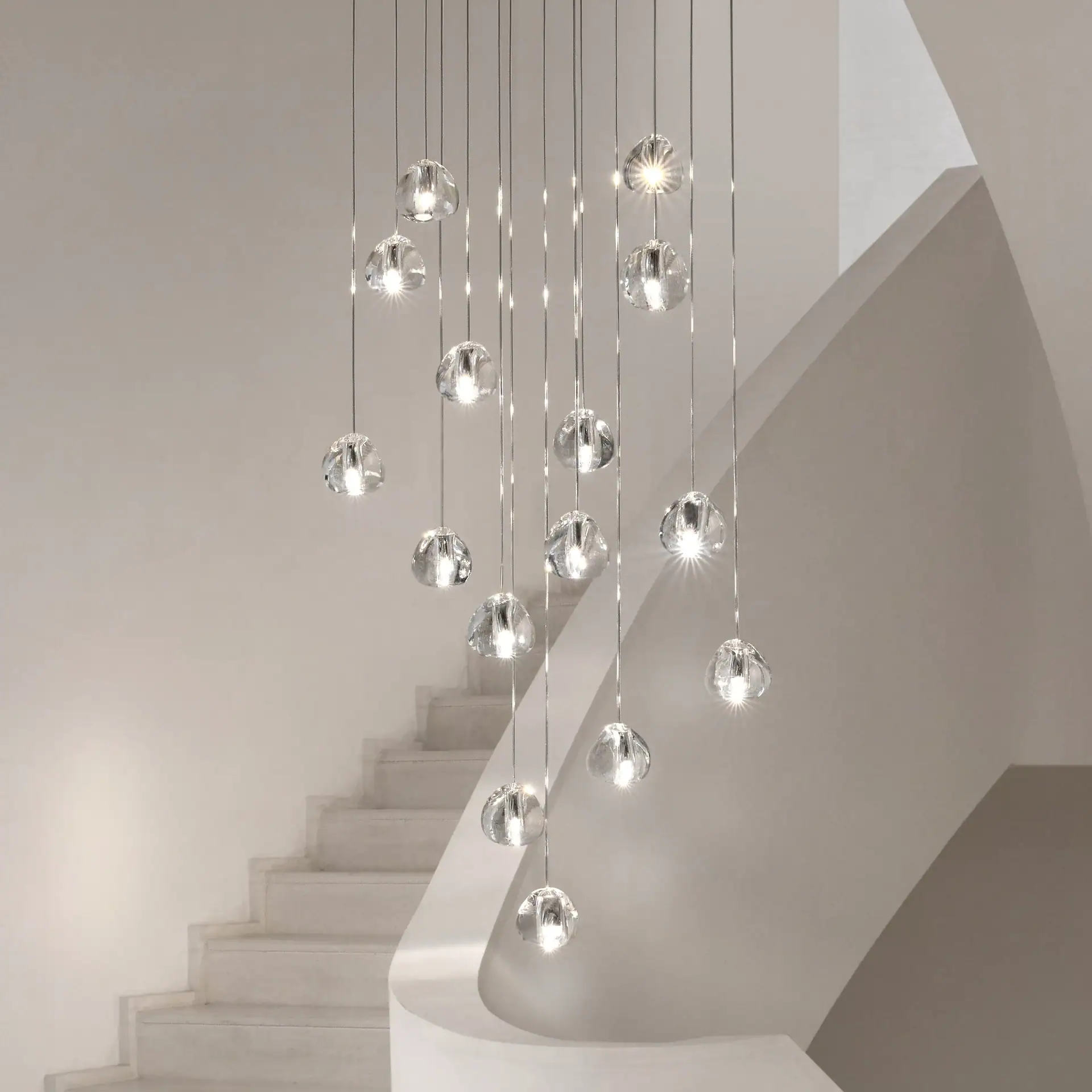 A modern chandelier with multiple clear glass globes hanging at different lengths, illuminated brightly, next to a light-colored curved staircase.