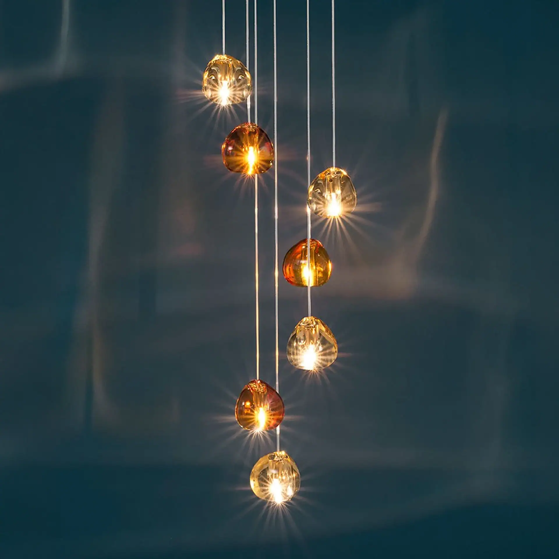 Eight amber and gold-toned glass pendant lights hanging vertically against a dark teal background, emitting a warm, starburst-like glow.