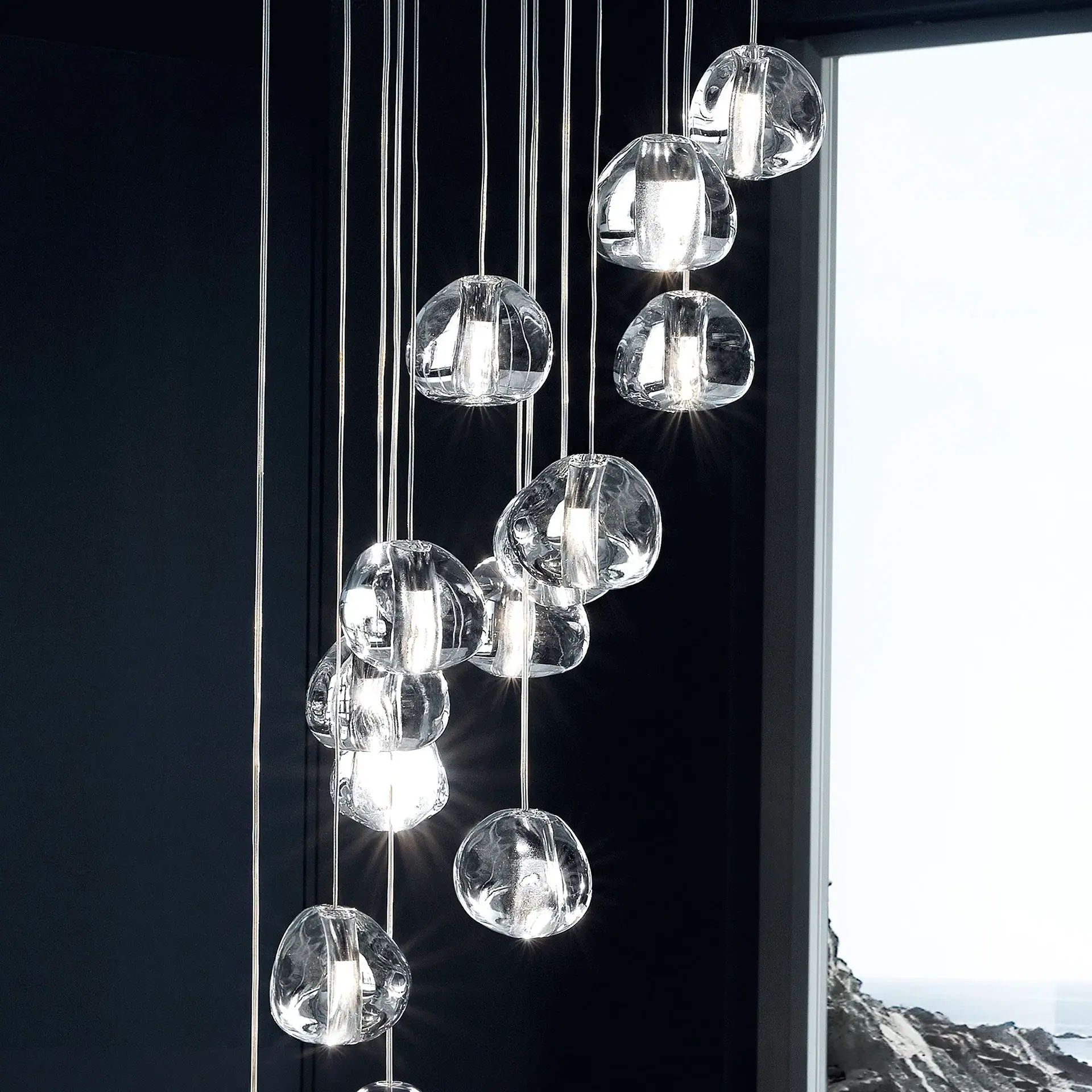 Modern chandelier with multiple clear, irregularly shaped glass pendants hanging at different lengths, illuminated against a dark wall and a window showing a rocky coastline.