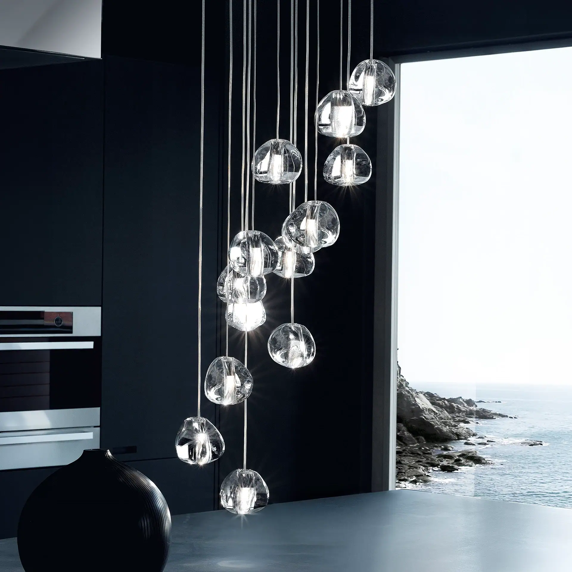 A modern kitchen with a cascading glass pendant chandelier overlooking a rocky coastline and ocean.