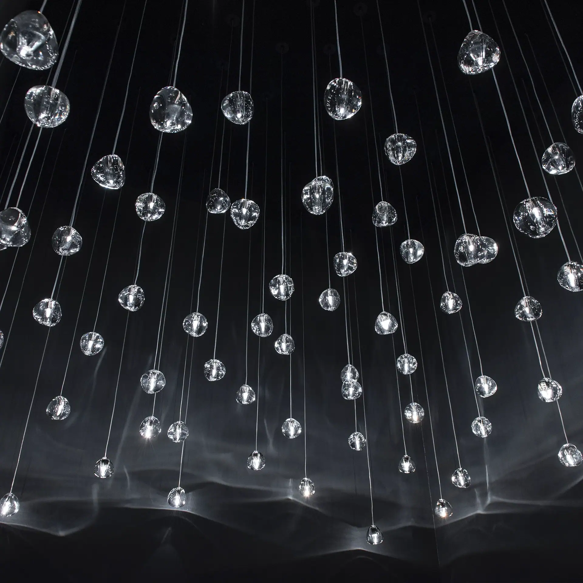 Numerous clear, irregularly shaped glass pendants hang from a dark ceiling, reflecting light and casting wavy patterns onto a surface below.