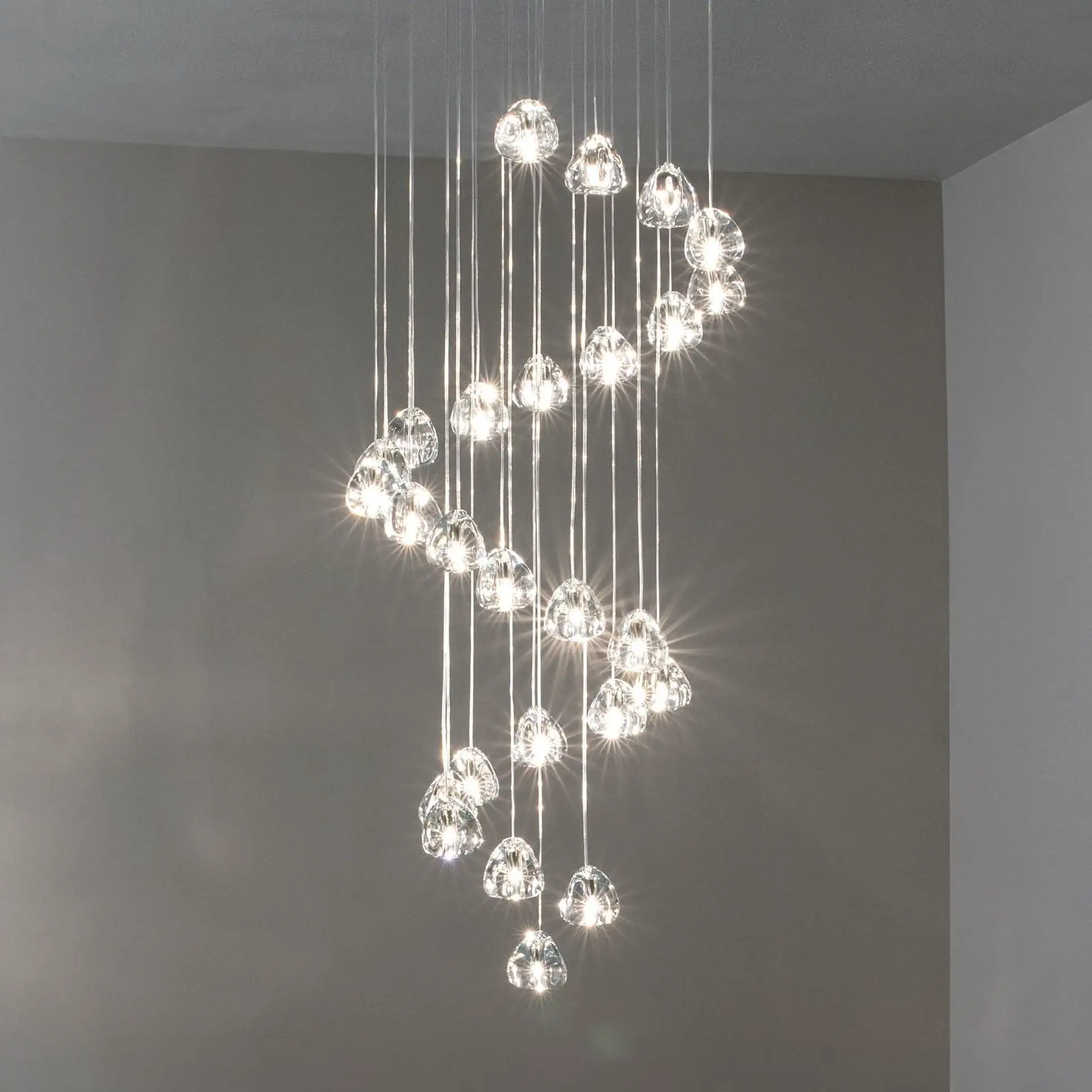 A modern spiral chandelier with many clear glass pendants, each emitting bright light, against a plain gray wall and ceiling.