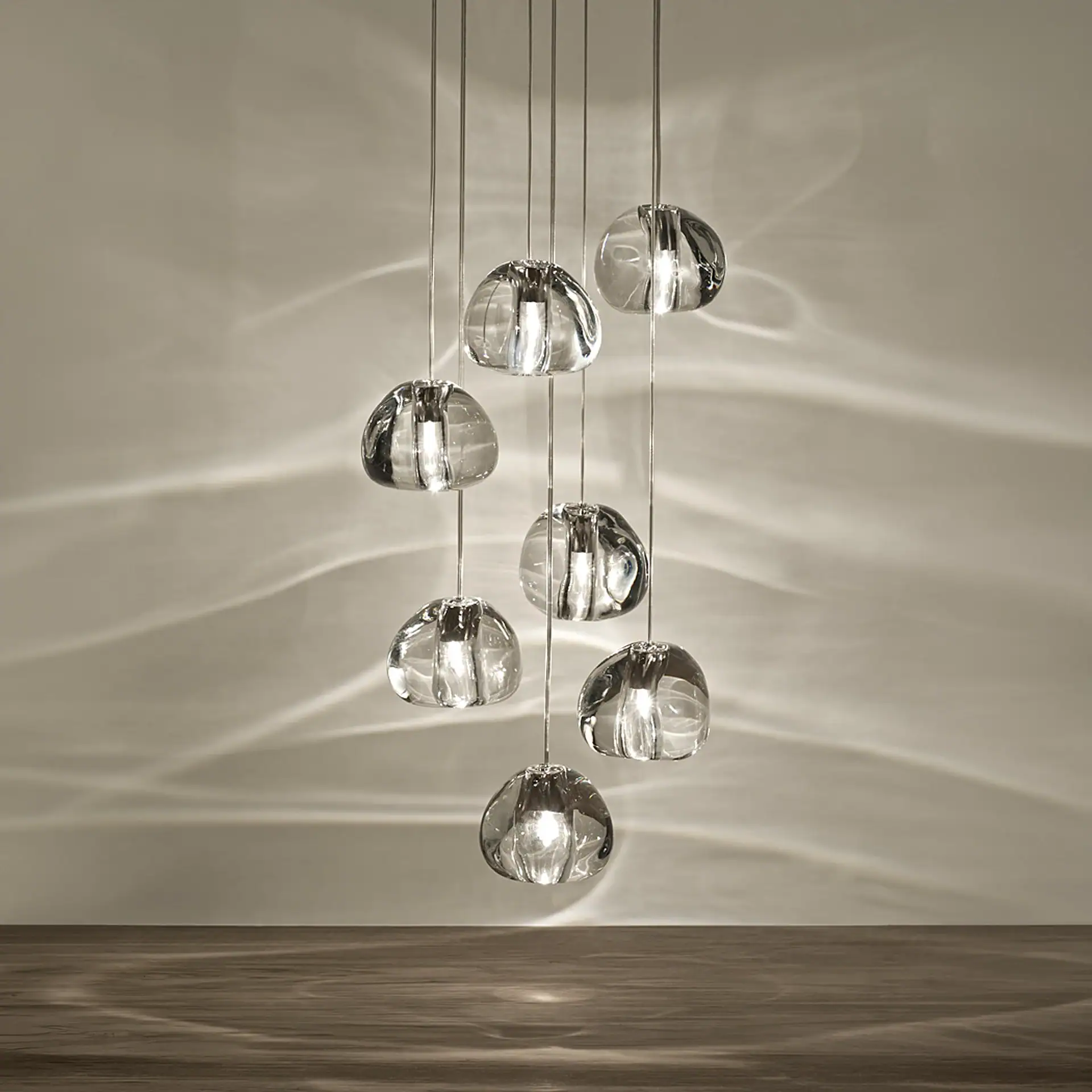 Eight clear, irregularly shaped glass pendant lights hanging at different heights, casting wavy light patterns on a light wall and dark wooden floor.