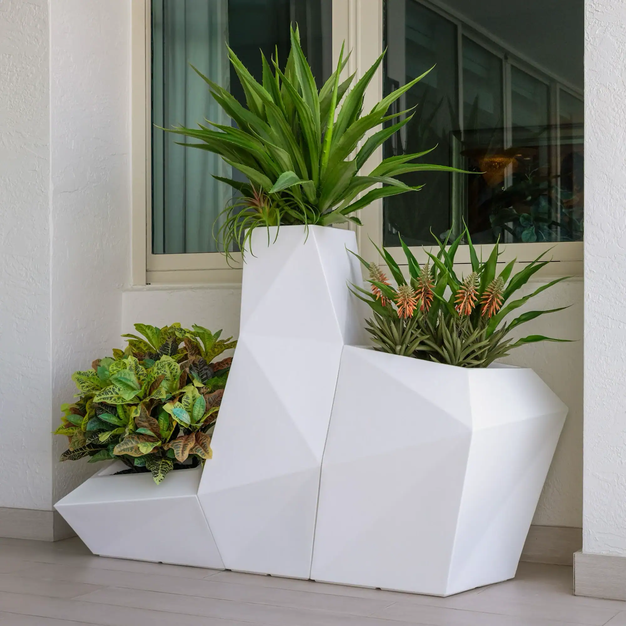 Modern geometric planter with greenery White geometric planter with various green plants outside near a window.