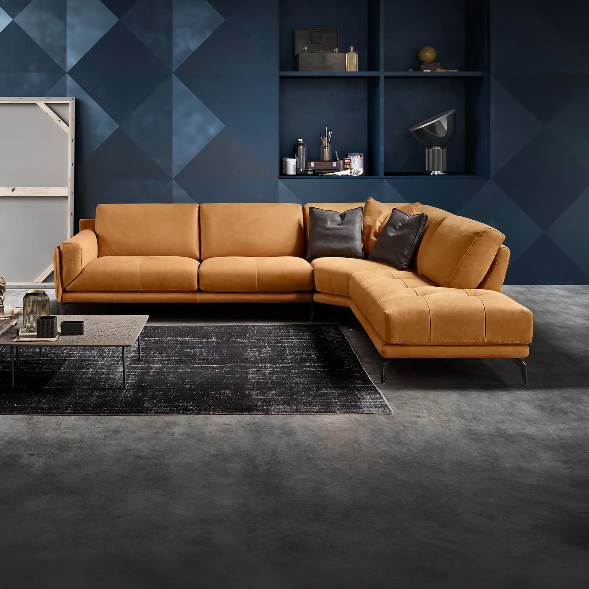 A modern living room with a large L-shaped light brown leather sectional sofa, a dark blue geometric wall with shelves, a distressed rug, and a gray coffee table.