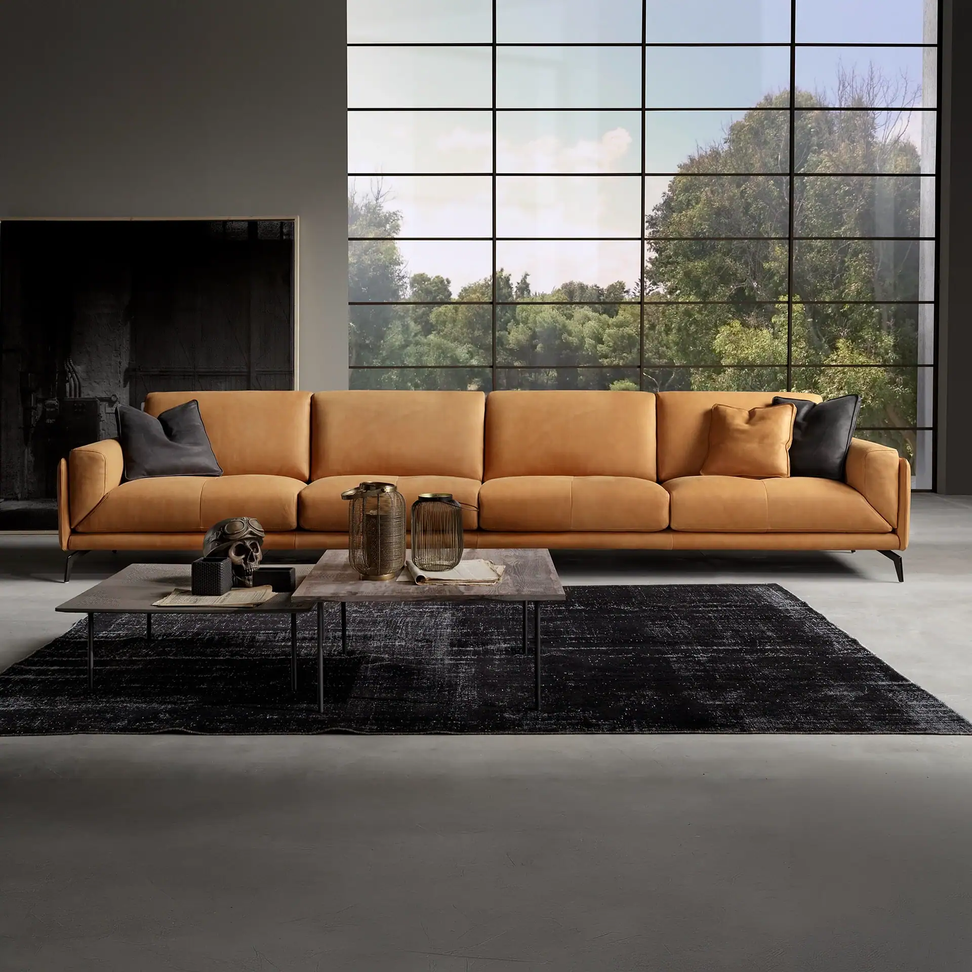 A modern living room with a long tan leather sofa, two coffee tables with decorative items, a black distressed rug, and a large grid window overlooking green trees.