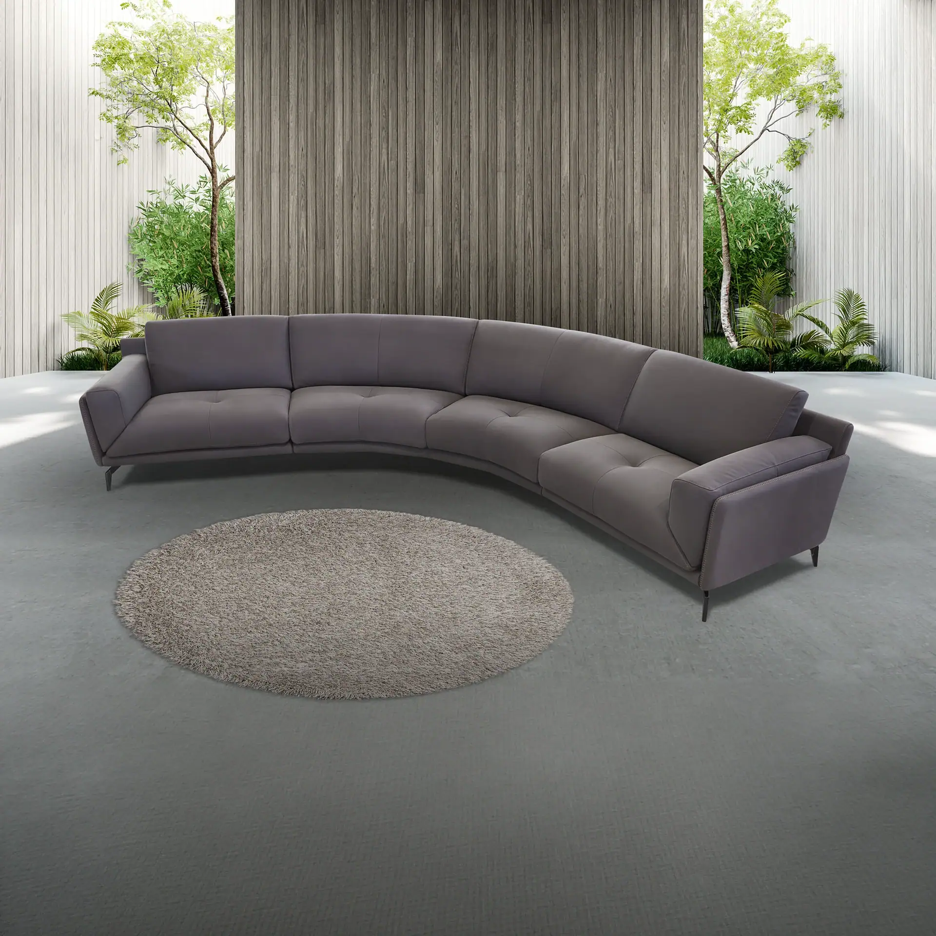 A charcoal gray curved sectional sofa on a concrete floor with a round shaggy rug. Behind it, wood-paneled walls reveal green trees and plants.