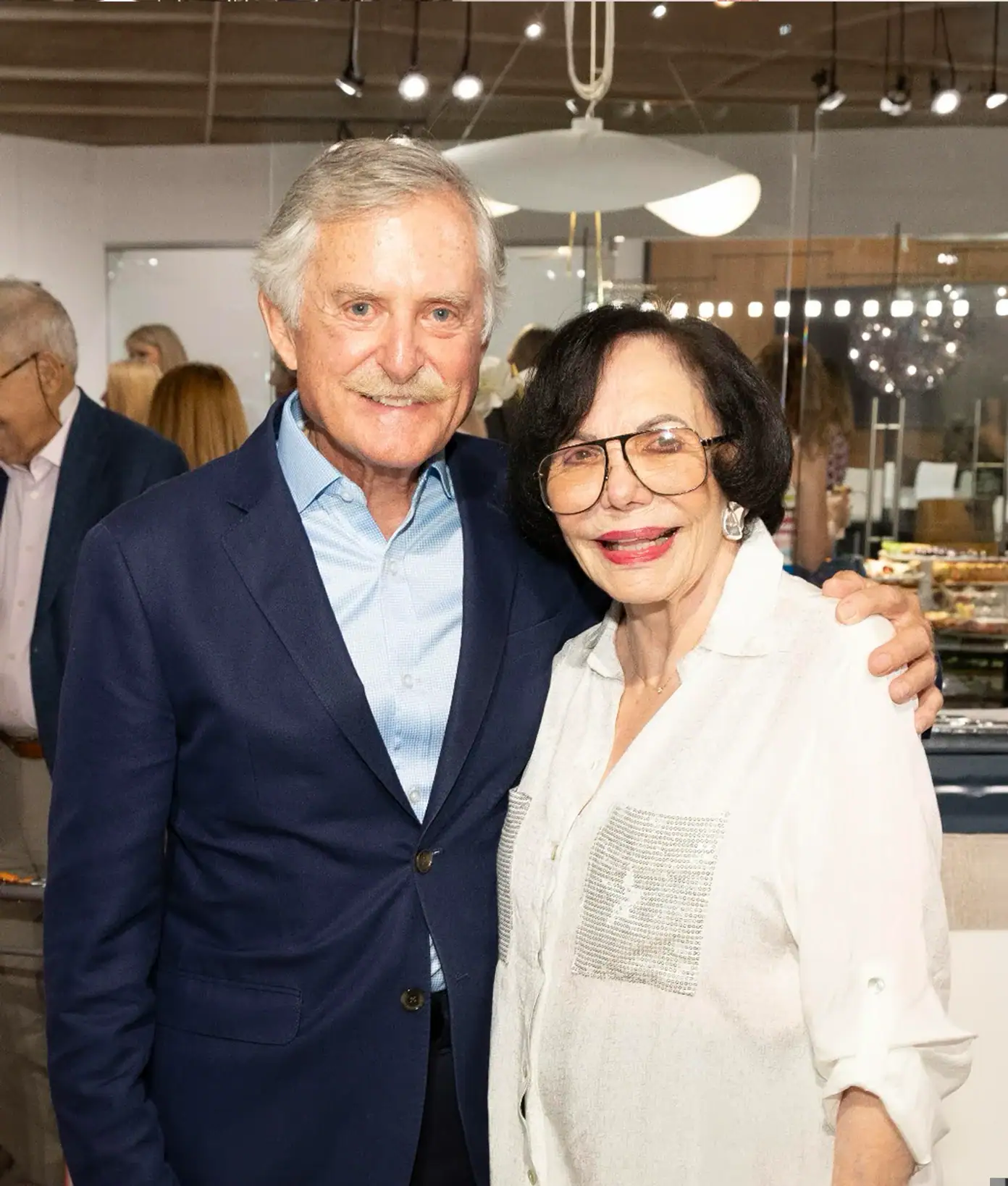 Smiling elderly man and woman at event, embracing and looking at camera.