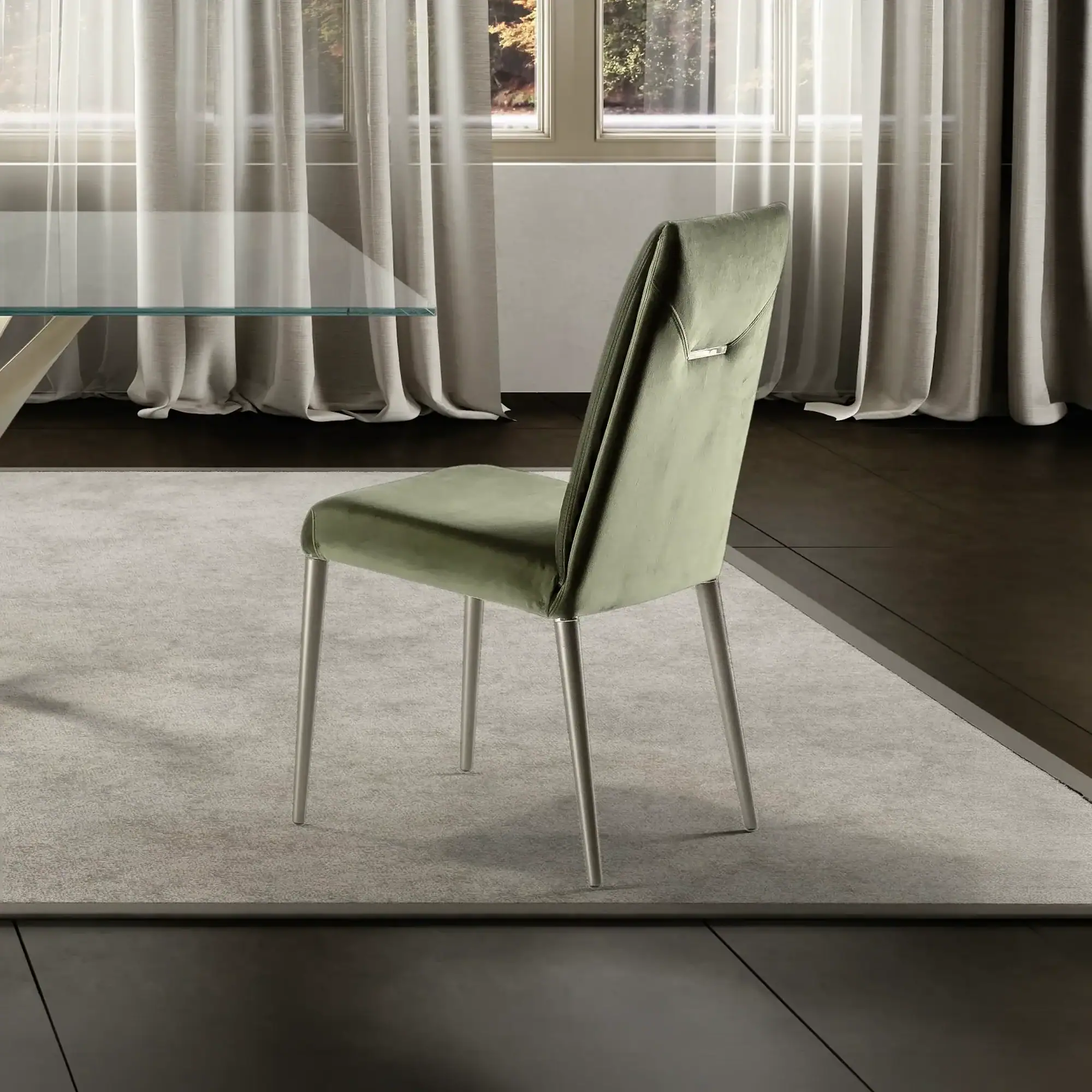 A modern olive green velvet dining chair with silver legs on a gray rug, with a glass table and sheer curtains in the background.