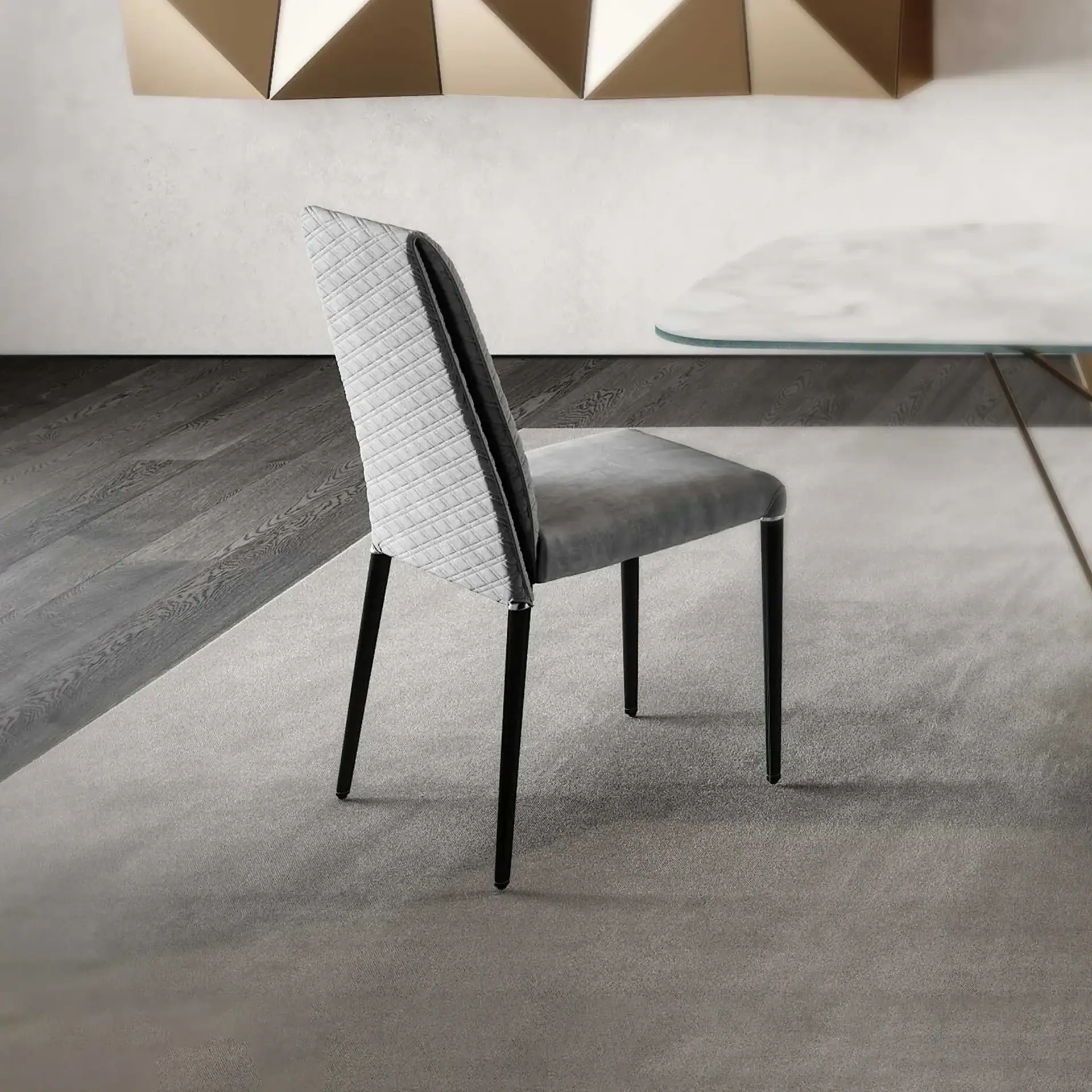 A modern grey dining chair with a quilted backrest on a light rug, next to a dining table and geometric wall art.