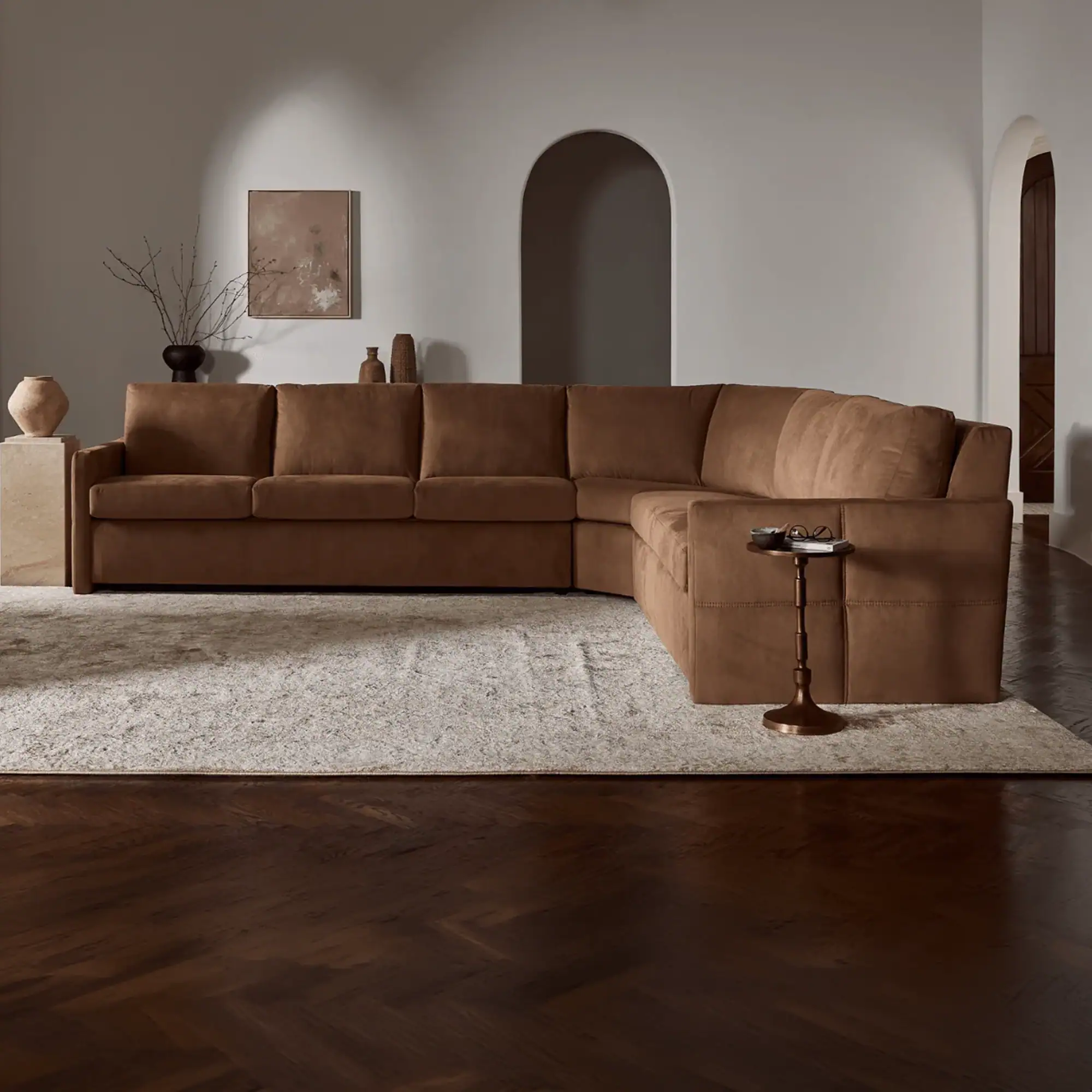 A warm brown L-shaped sectional sofa sits on a light rug in a modern living room with dark herringbone floors and arched doorways. Decor includes abstract art, vases with branches, and a small side table.