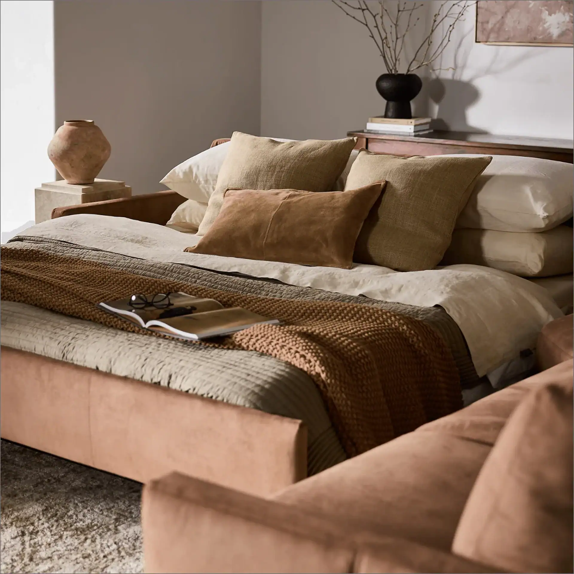 A daybed with neutral bedding, layered blankets, an open book, and glasses, flanked by a decorative vase and a side table with branches and books.