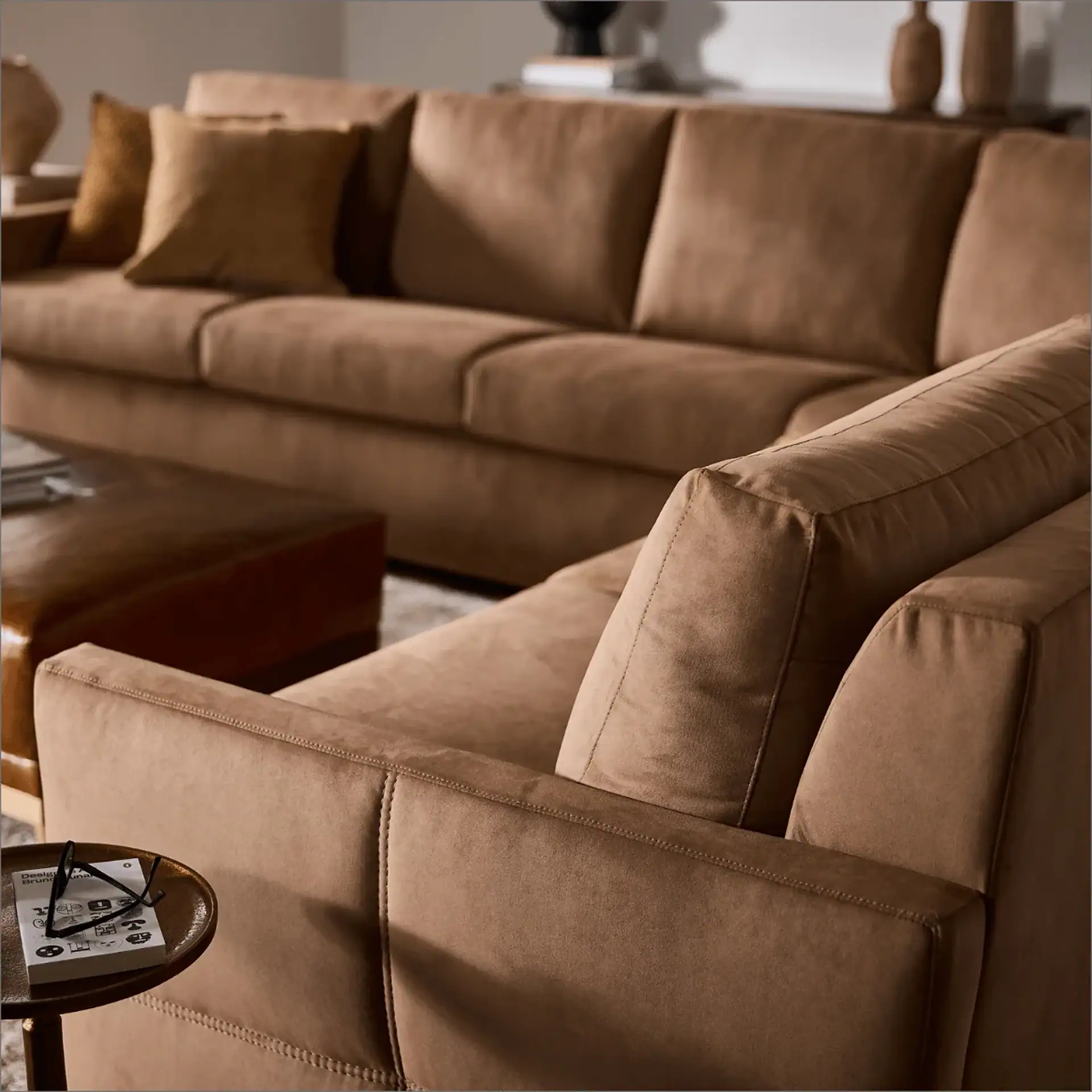 A large, L-shaped brown sectional sofa with throw pillows in a modern living room. A small side table holds a book and glasses in the foreground.