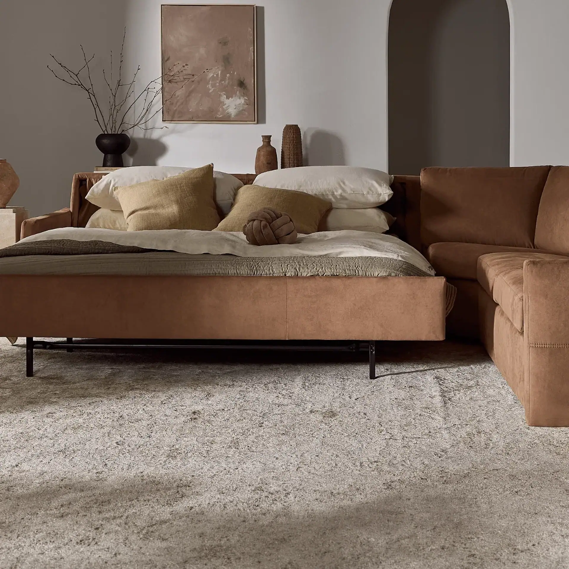 A modern living room featuring a brown L-shaped sofa converted into a bed, adorned with neutral pillows and blankets, set on a textured gray rug. Abstract art and ceramic vases decorate the background.
