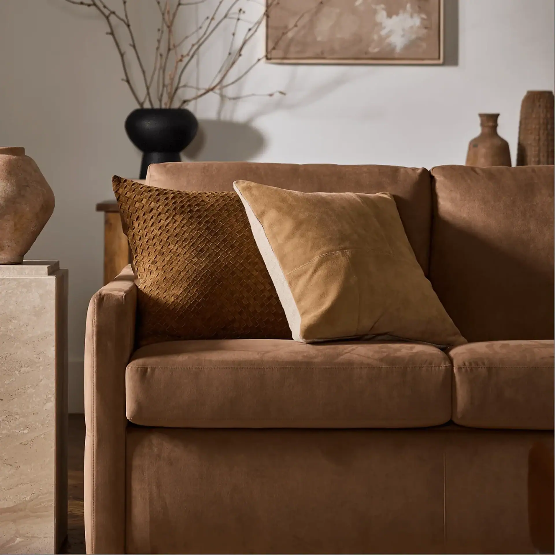 A brown sofa with two decorative pillows: one dark brown with a woven texture, and one light tan with a smooth finish. Various rustic vases and a black vase with branches are in the background.