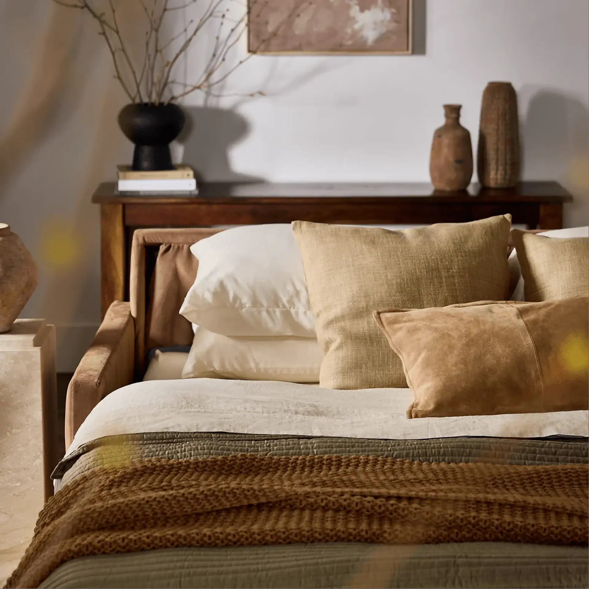A bed with layered white, olive-green, and brown bedding, and multiple pillows. A wooden console table behind it holds vases and books, with an abstract artwork on the wall.