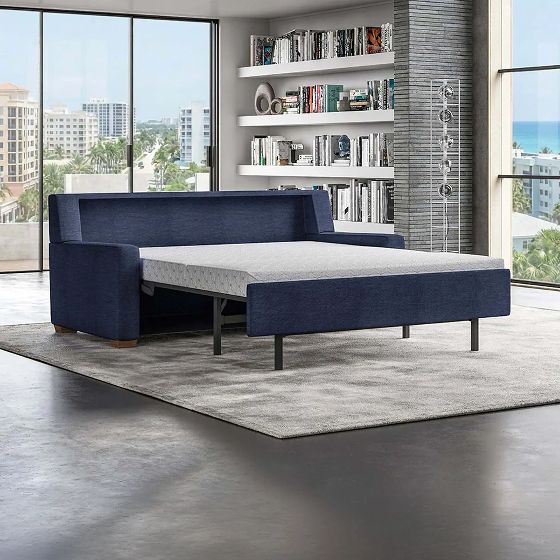 A navy blue sofa bed extended into a bed in a modern room with large windows overlooking a city and ocean. A bookshelf is visible in the background.