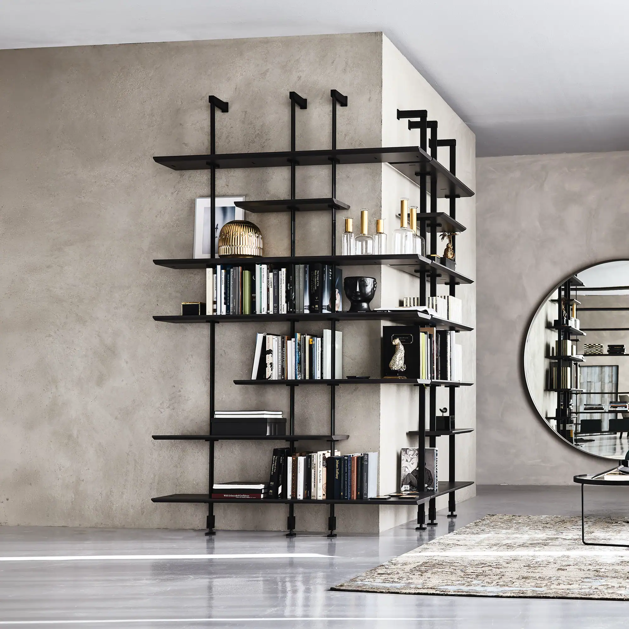 A modern black modular bookshelf filled with books and decor, against a gray wall, with a round mirror and patterned rug on a polished concrete floor.