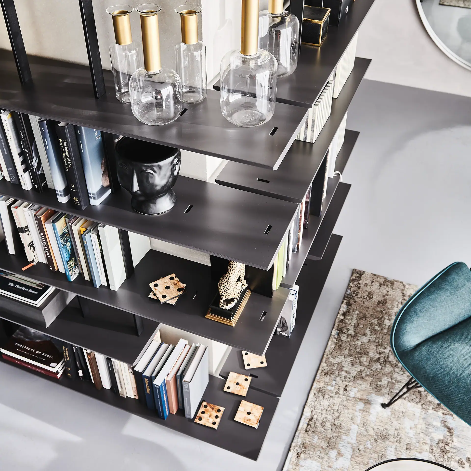 A dark grey modular bookshelf filled with books, glass vases, and decorative items, next to a rug and a teal chair.