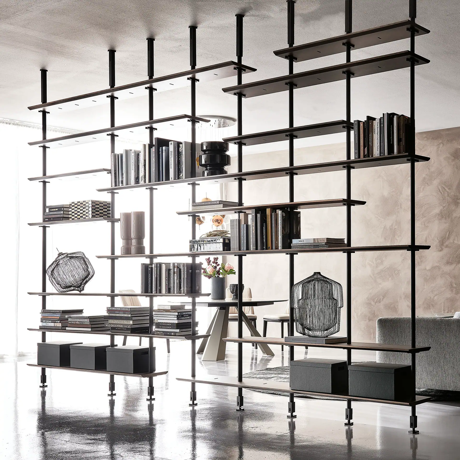 A modern, dark wood and metal shelving unit acting as a room divider, filled with books, decorative objects, and storage boxes in a bright, contemporary interior.