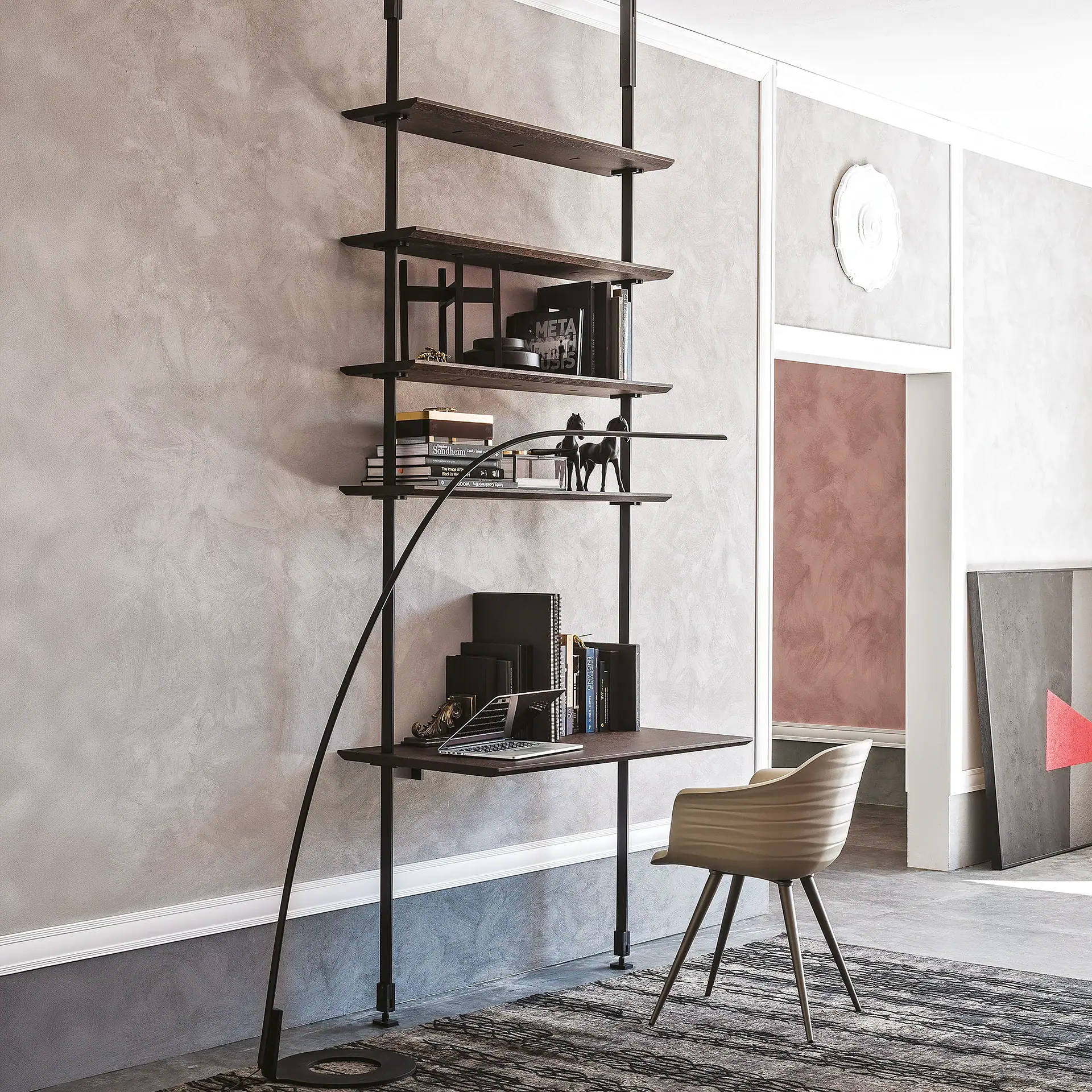 A modern wall-mounted desk and shelving unit with a laptop and books, a light beige chair, and a curved floor lamp in a room with textured gray walls and a patterned rug.