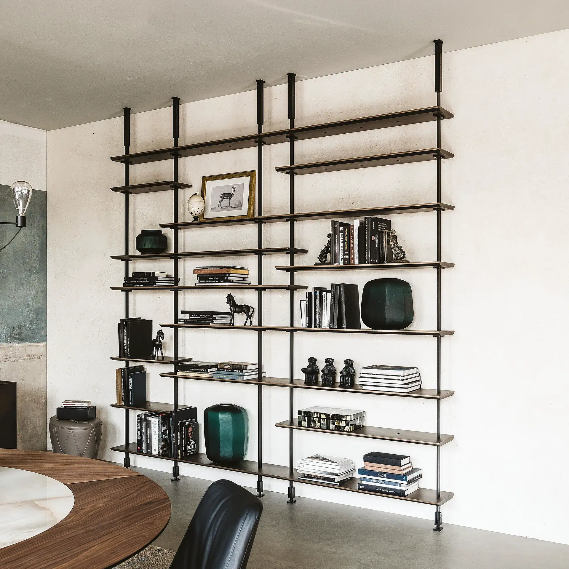 A modern living room with a large, dark wood and metal modular shelving unit filled with books and decorative items against a white wall. A round dining table and black chair are in the foreground.