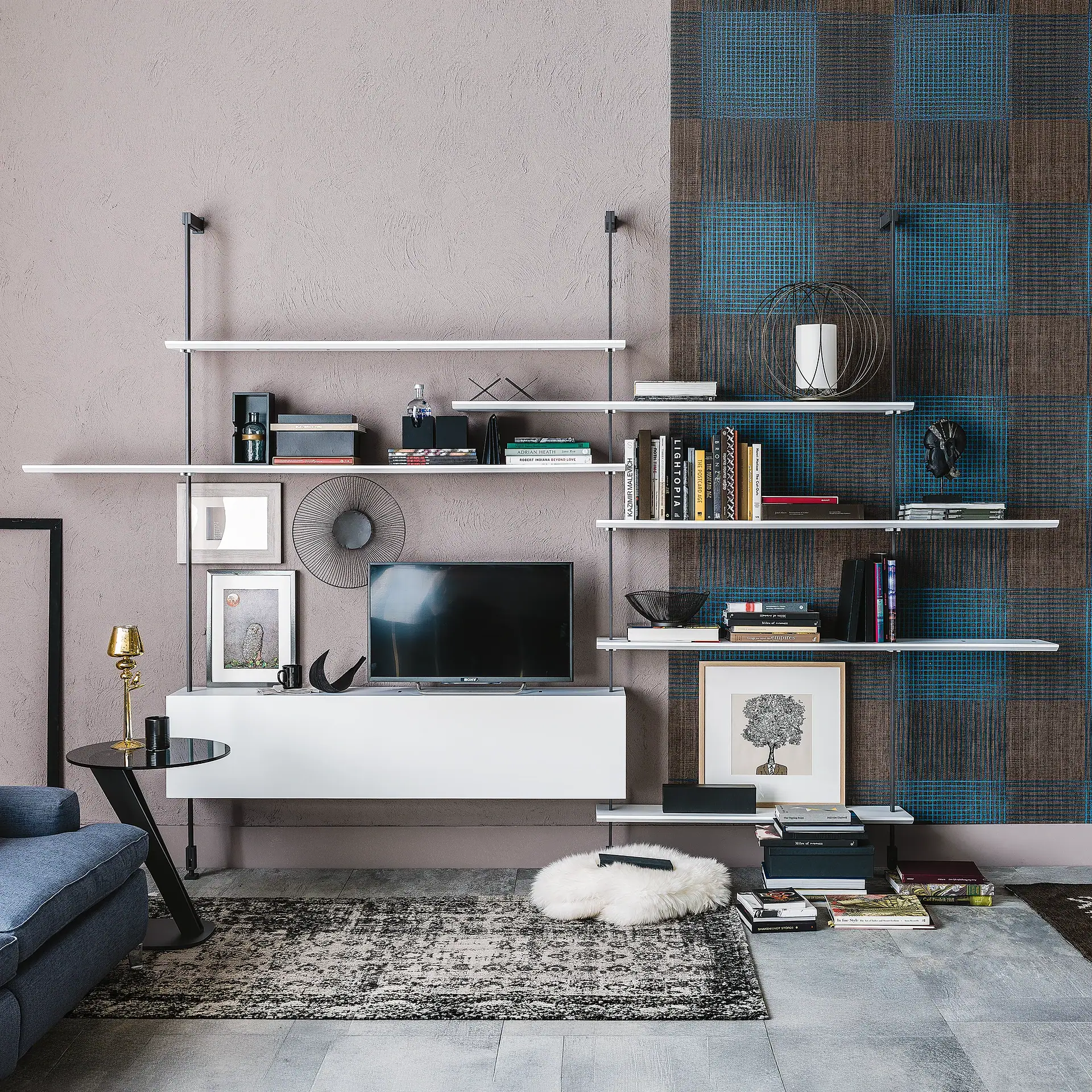 A modern living room with a white wall-mounted shelving unit displaying books and decor, a television, and a blue sofa on a patterned rug.