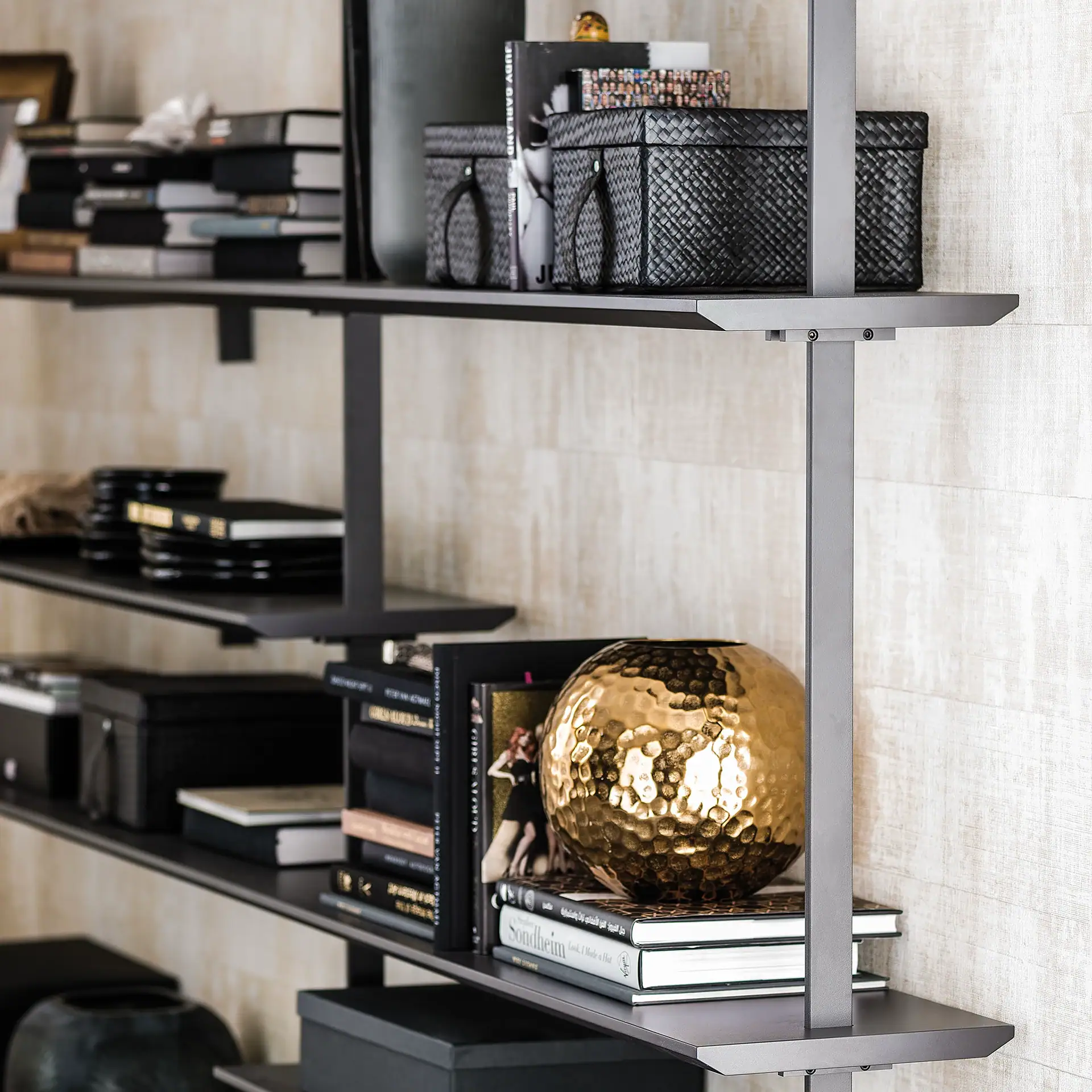 A modern dark gray shelving unit filled with books, black woven baskets, and a hammered gold sphere against a light textured wall.