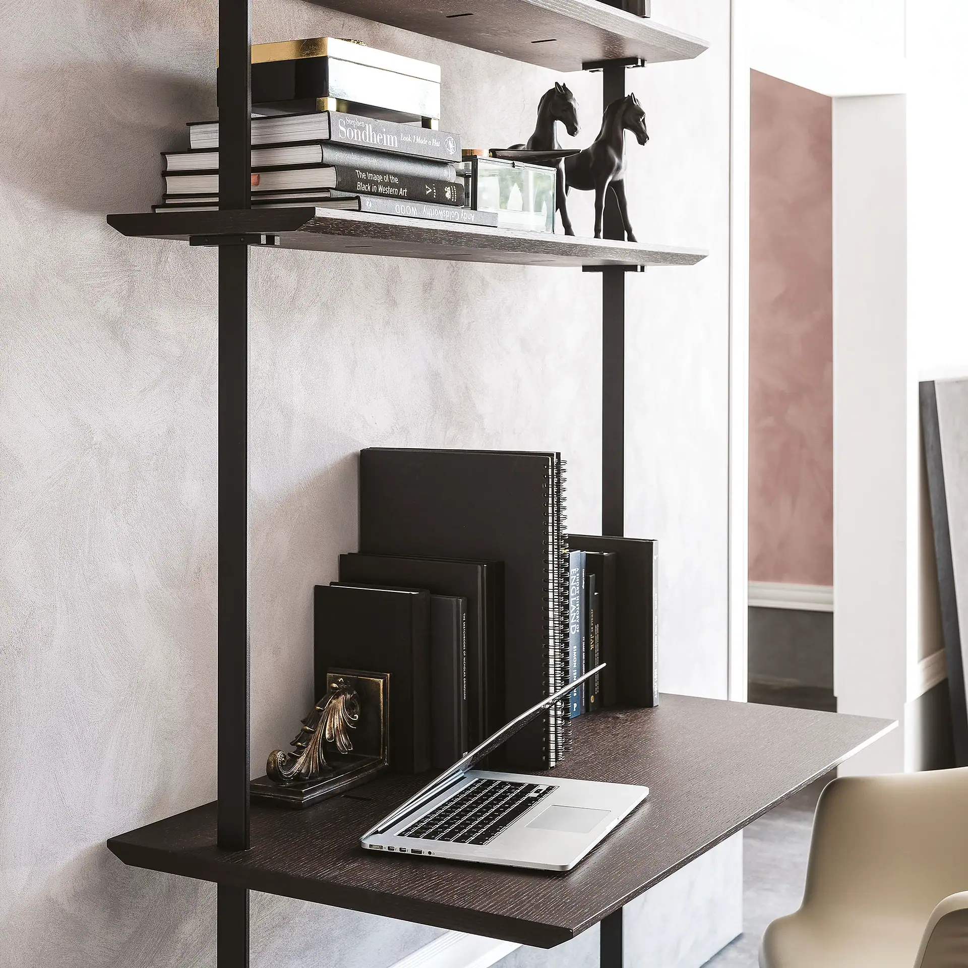 A modern dark wood shelving unit with a desk, books, decorative horse figurines, and a silver laptop on a textured light wall.